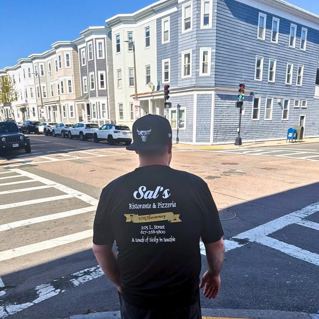 A man wearing a black Chicago Bulls hat and a black T-shirt with white text on the back stands at a crosswalk in a city neighborhood with multi-story houses and cars parked along the street.