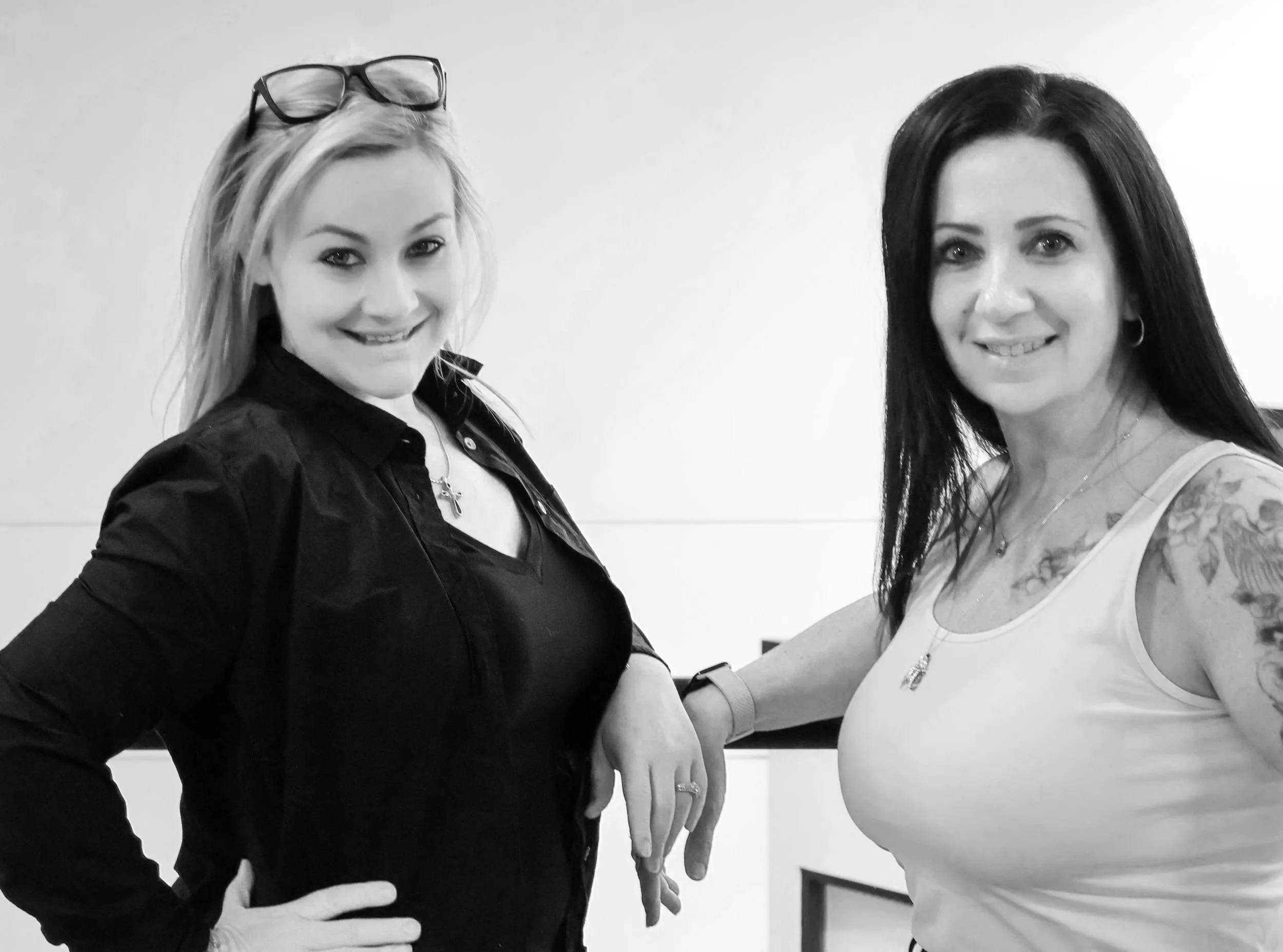 Two women standing close together, smiling, with one woman wearing glasses on her head and the other wearing earrings and a necklace. The woman on the right has a floral pattern tattoo on her upper arm.