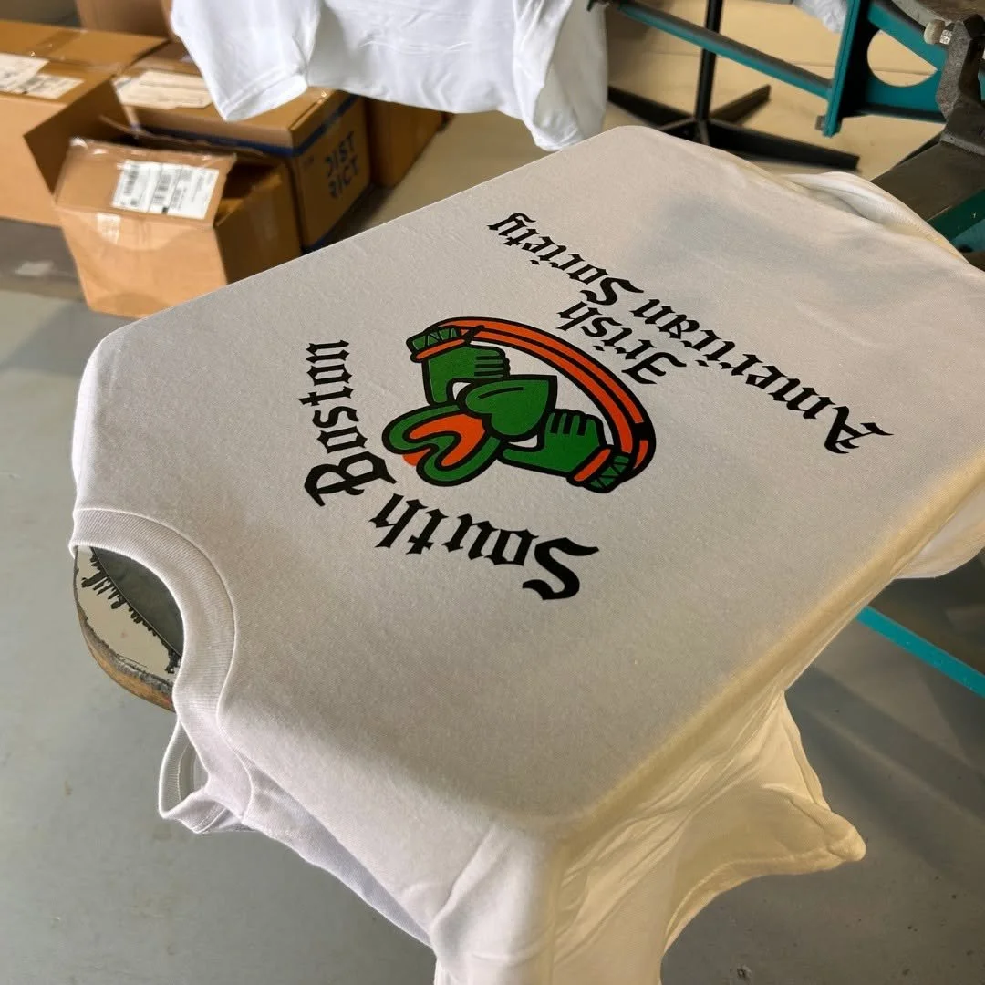 A white T-shirt with a logo that says "Soxy Bush" and depicts green boxing gloves, laid on a table in a room with cardboard boxes and a cleaning cloth in the background.