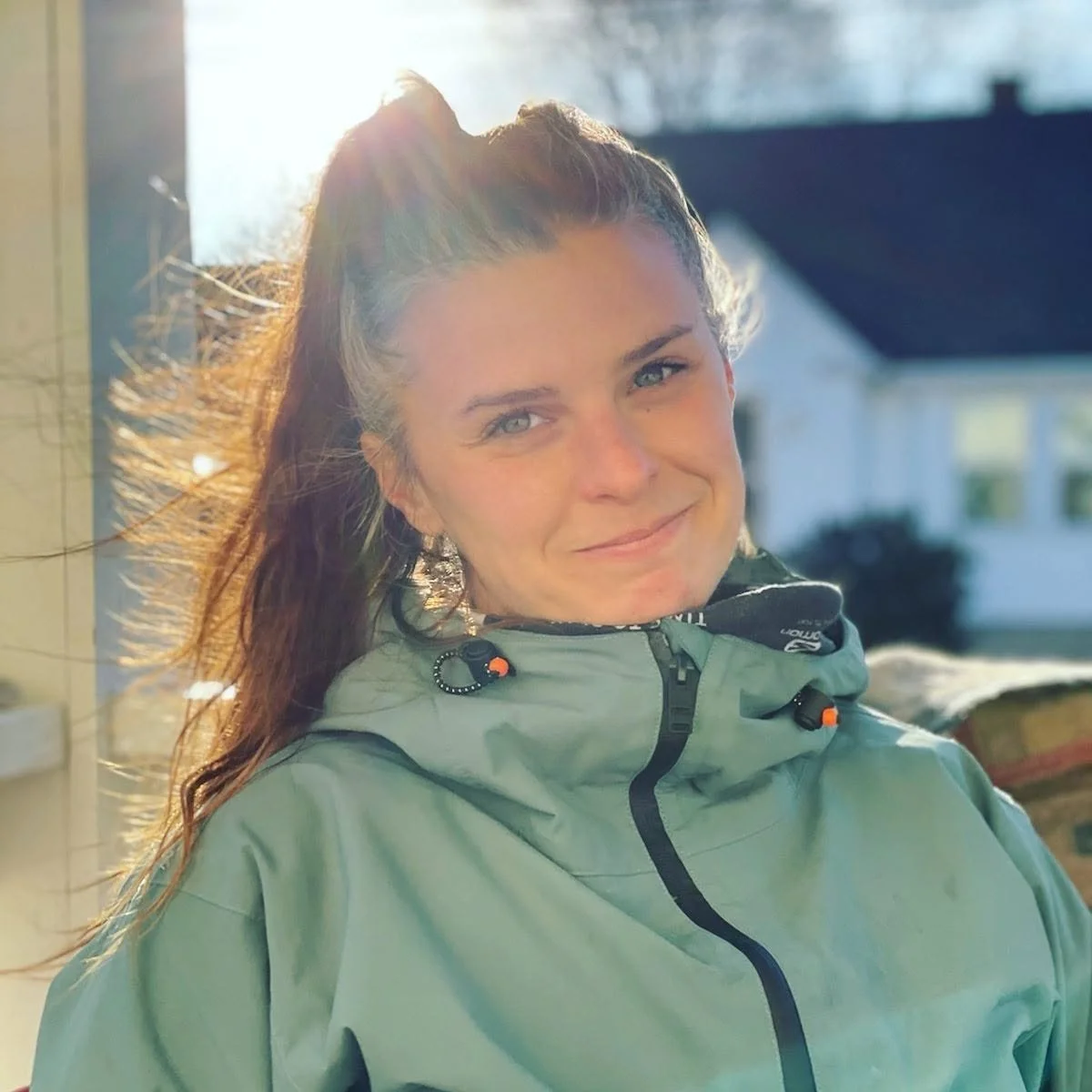 Smiling woman with a ponytail wearing a green outdoor jacket in warm sunlight