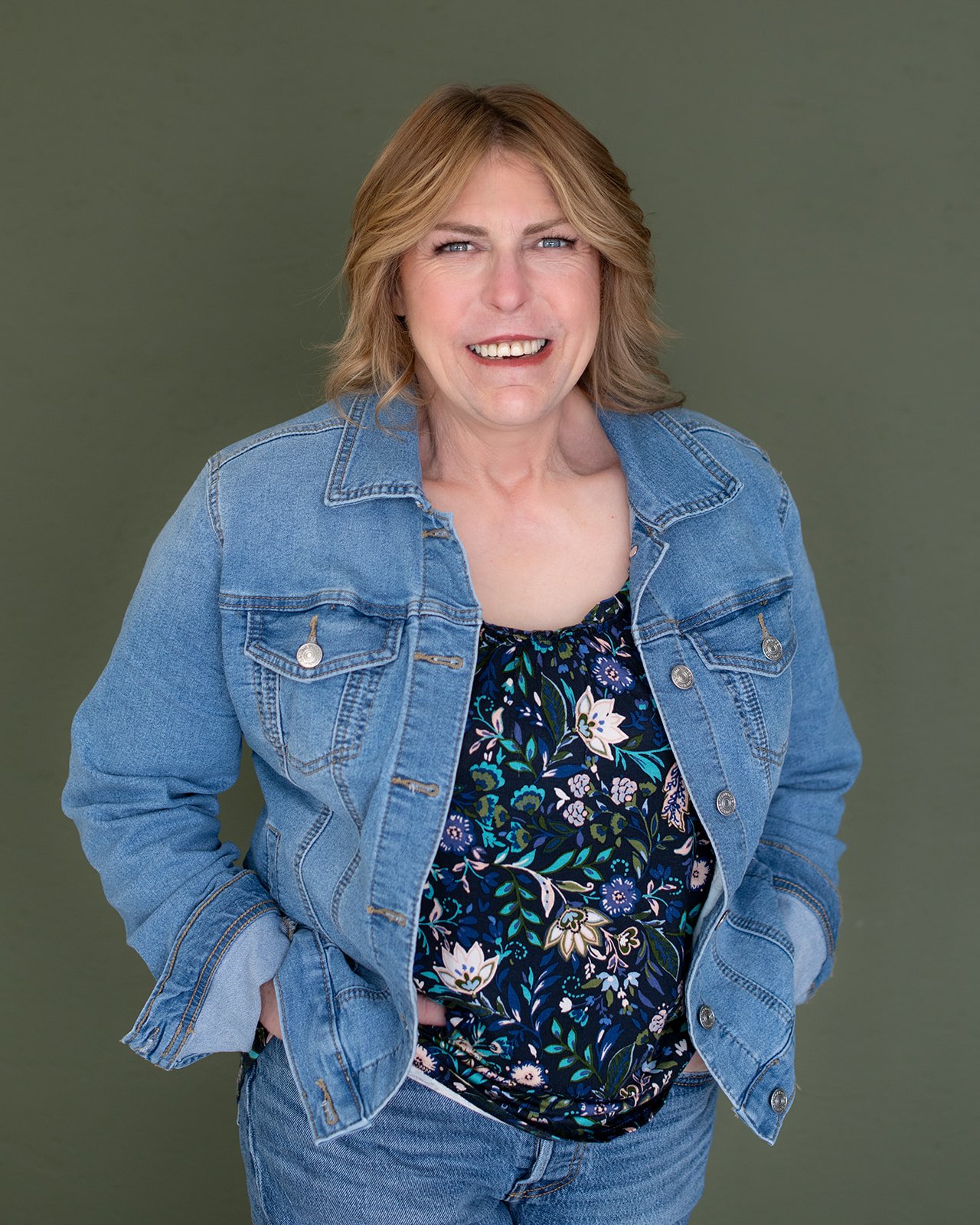 Portrait of a middle-aged woman with blonde hair, wearing a denim jacket over a floral top, smiling and standing against a green background.