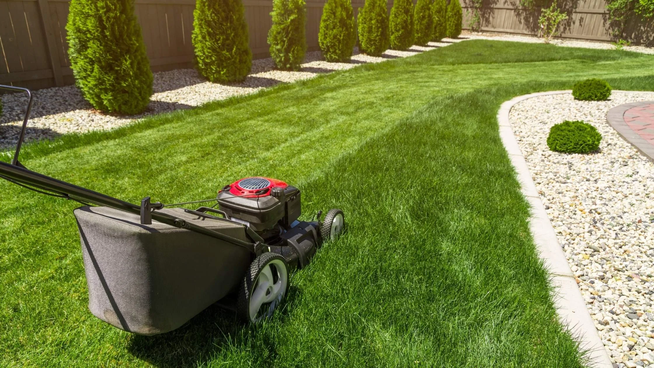 A lawn mower on a well-maintained grassy backyard with trimmed bushes and a decorative gravel border.