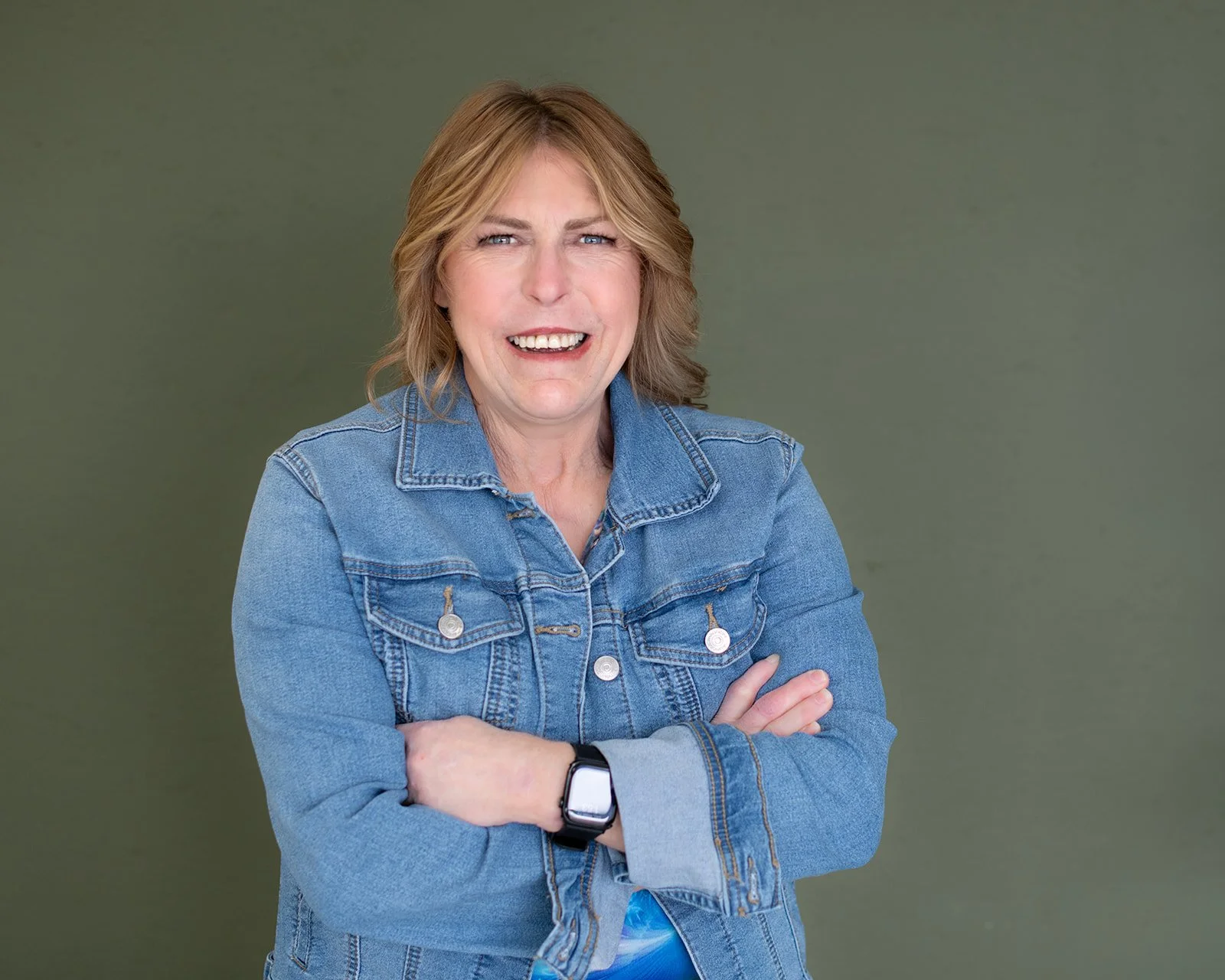 A woman with blonde hair wearing a blue denim jacket, smiling with arms crossed, standing against a green background.