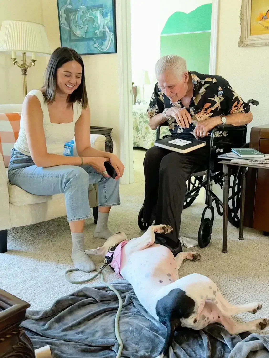 A young woman and an elderly man in a wheelchair sitting in a living room, smiling and looking at a dog lying on its back on the floor. The dog is pink and white, with a harness, and the room is decorated with paintings and a lamp.