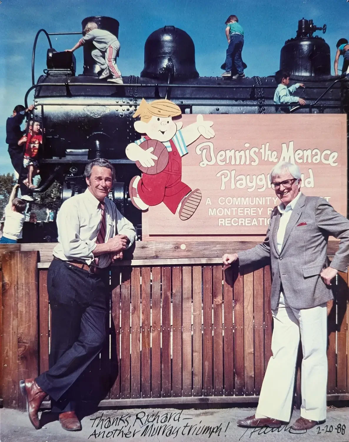 Two men standing in front of a sign for Dennis the Menace Playground, with children playing on a large black steam engine train behind them. The sign features a cartoon of Dennis the Menace. The photograph is dated February 20, 1988, and the caption thanks Richard for another Murray triumph.