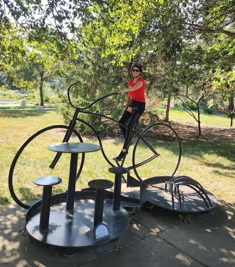 Person sitting on a bicycle-shaped sculpture in a park, smiling, wearing sunglasses and a red shirt.