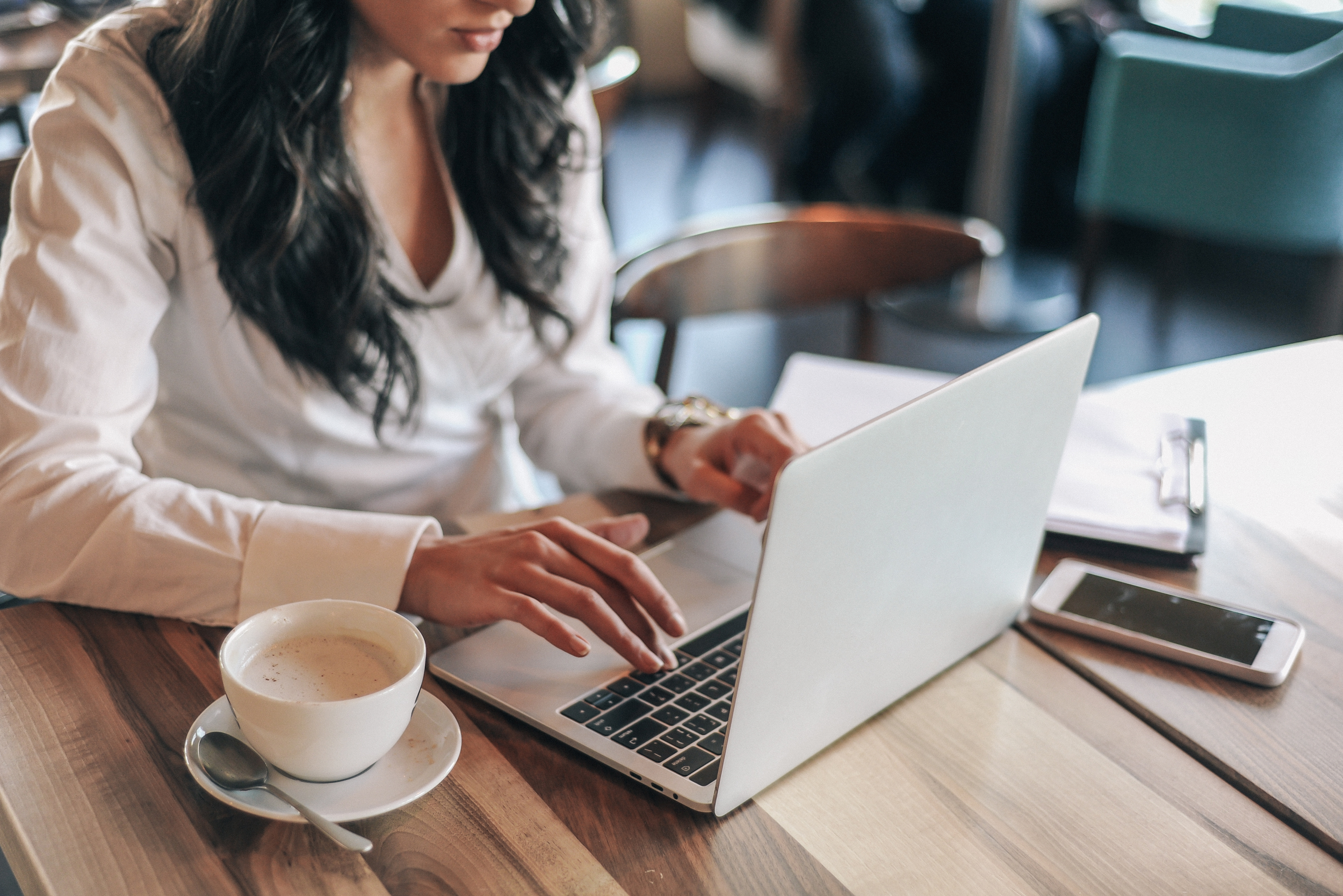 Woman in a white shirt working on a laptop at a wooden table in a cafe, with a cup of coffee and a smartphone nearby.
