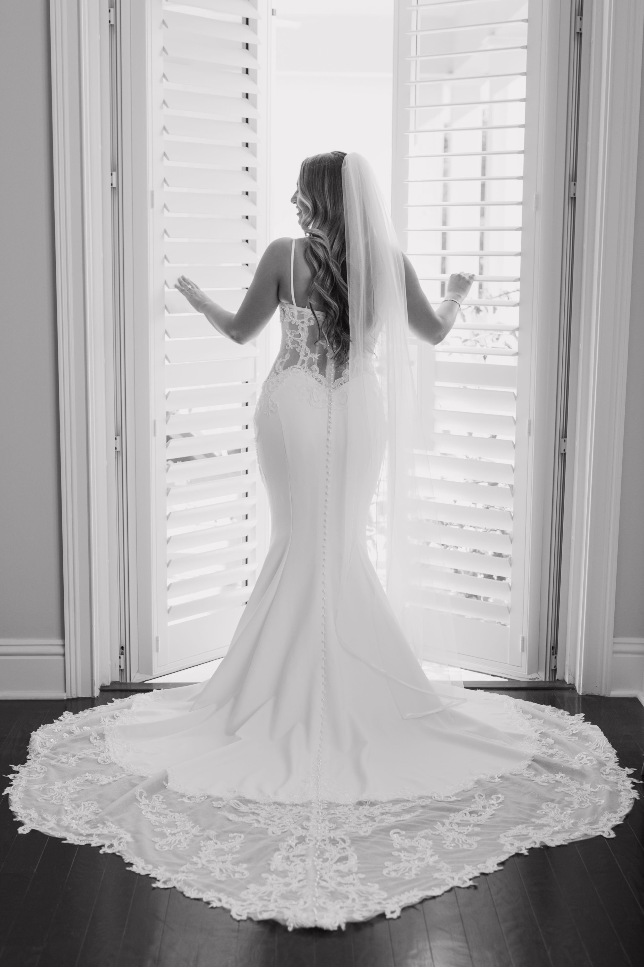 Bride in elegant wedding dress with lace train stands in front of open shutters in Palm Beach, FL | Shelby Eve Photography