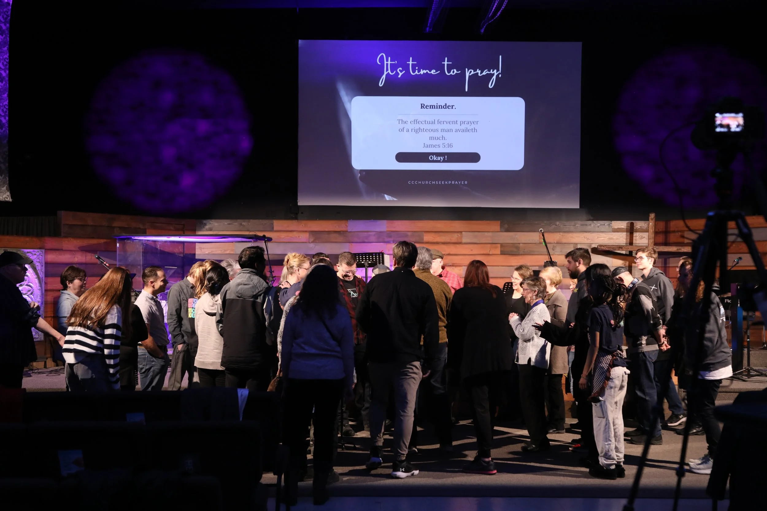 A group of people gathered in a church or auditorium with a large screen displaying a prayer reminder behind them.