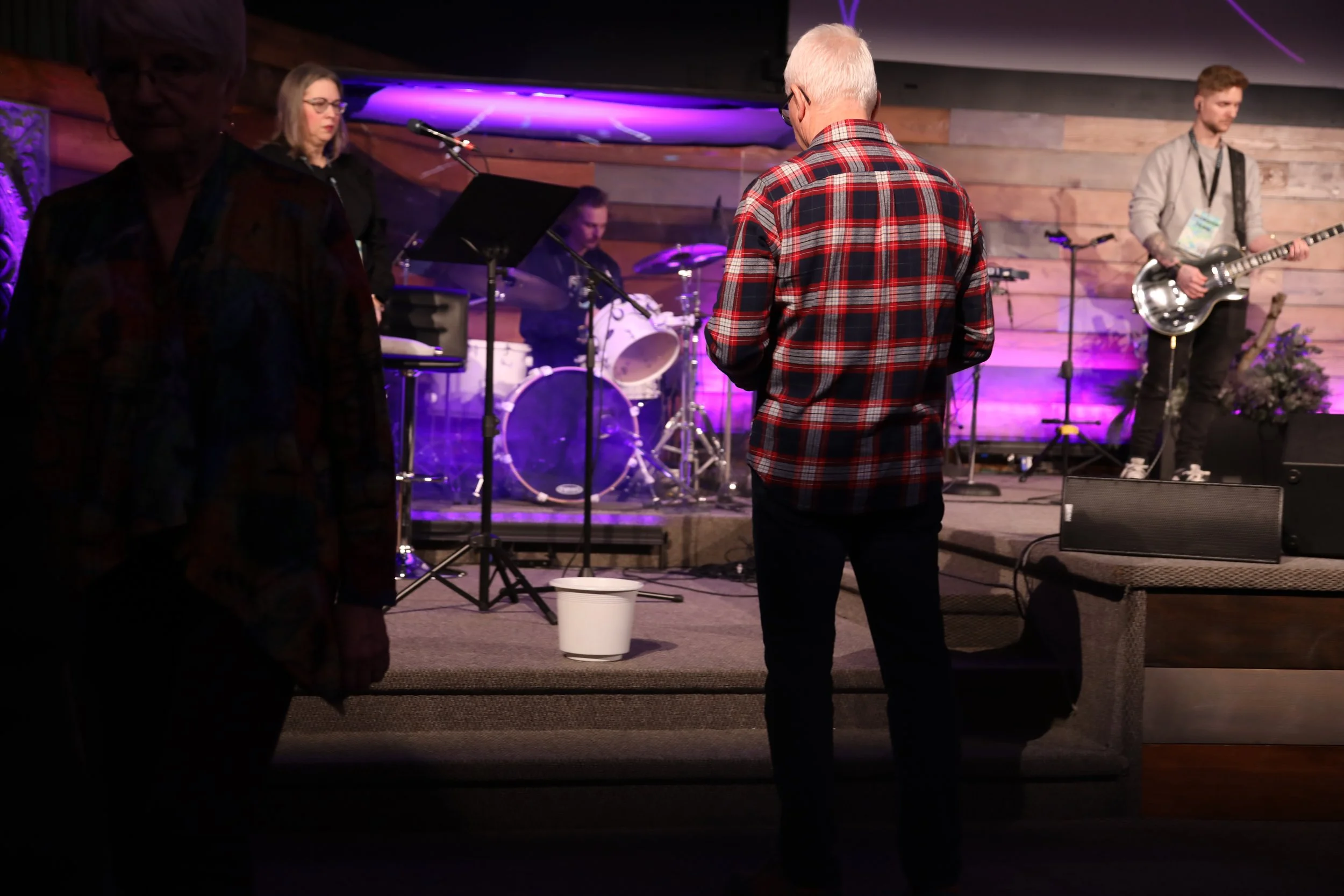 People watching a band perform on stage in a dimly lit venue with purple lighting. One person in a red plaid shirt stands in the foreground, facing the band, with others nearby.