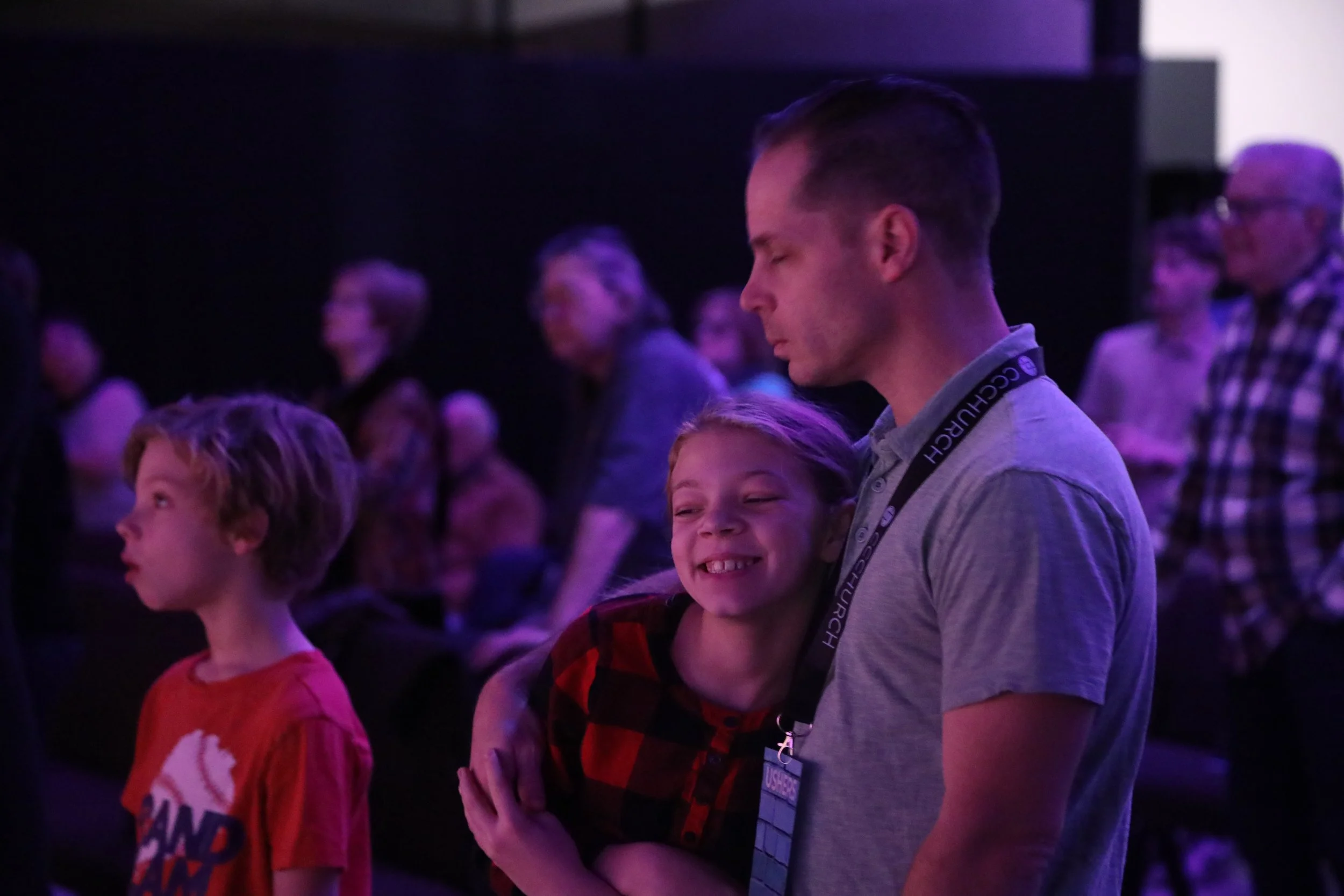 A man and a young girl are embracing at an event with other people in the background. The girl is smiling widely and the man has his eyes closed. The scene is dimly lit with purple and pink lighting.