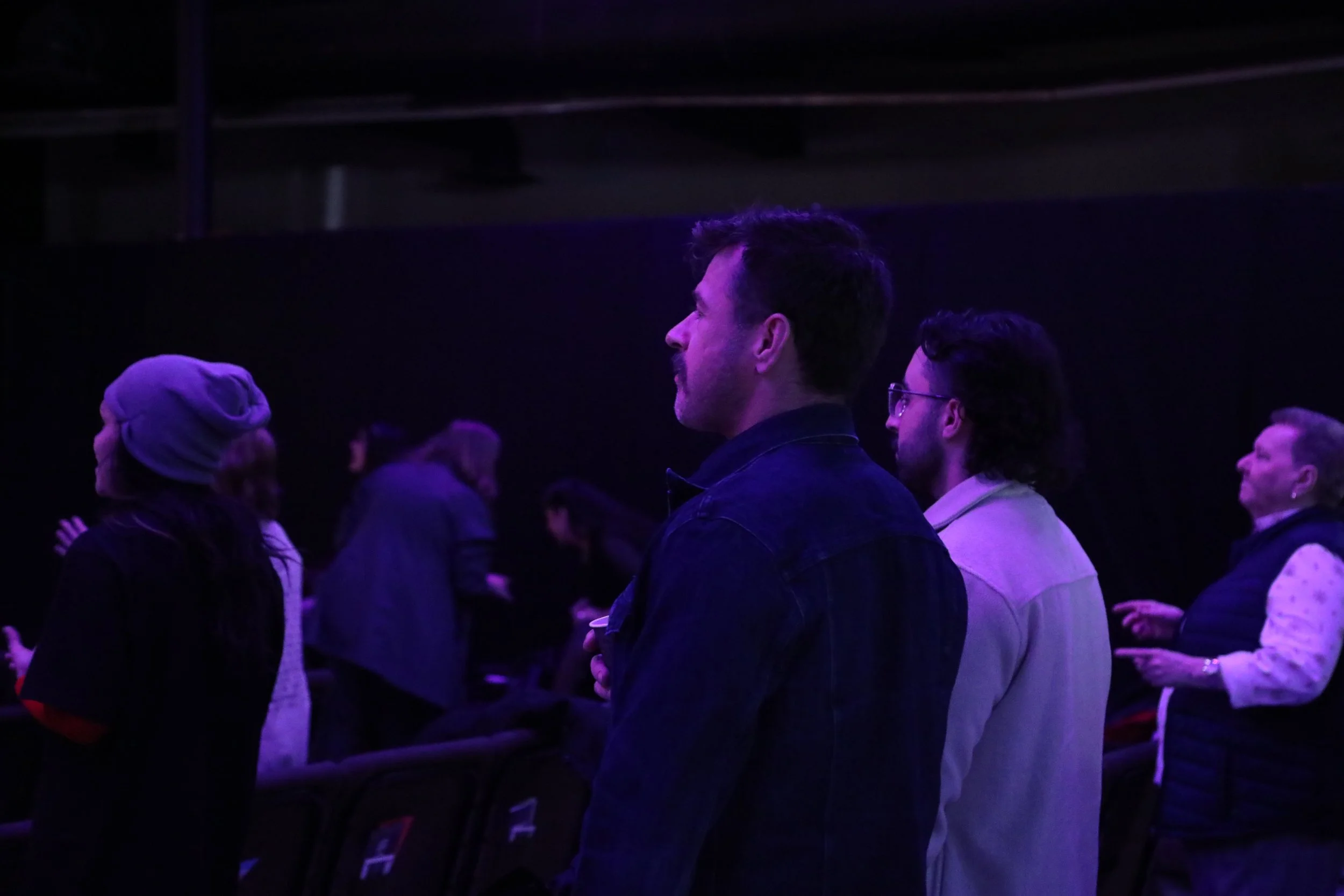 People standing in a dark theater or concert venue, observing or waiting, illuminated by purple lighting.