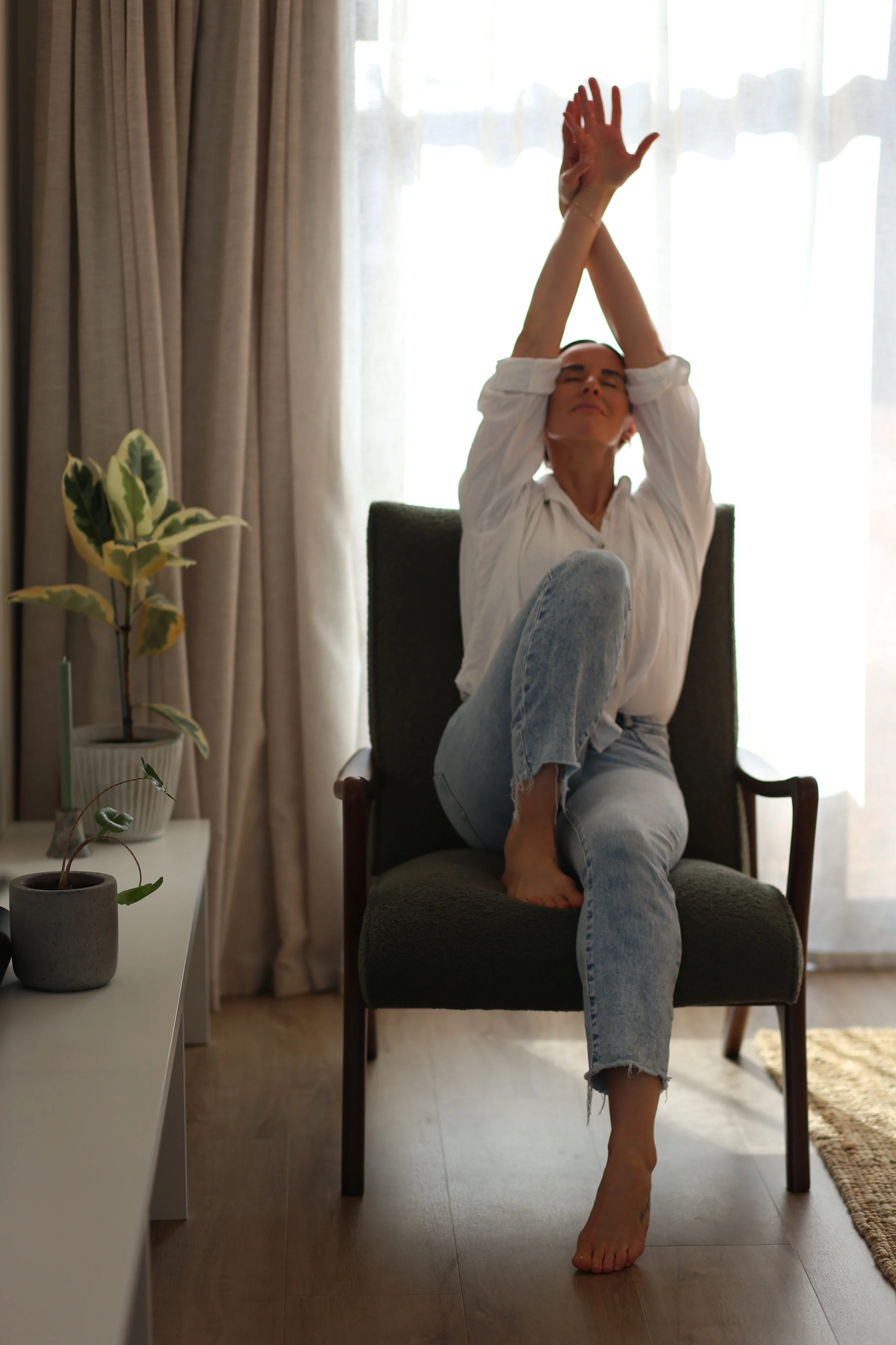 Holistic Wellness Coach - Alex Ballagh - is sitting on a green chair and stretching her arms up above her head in a yoga stretch.