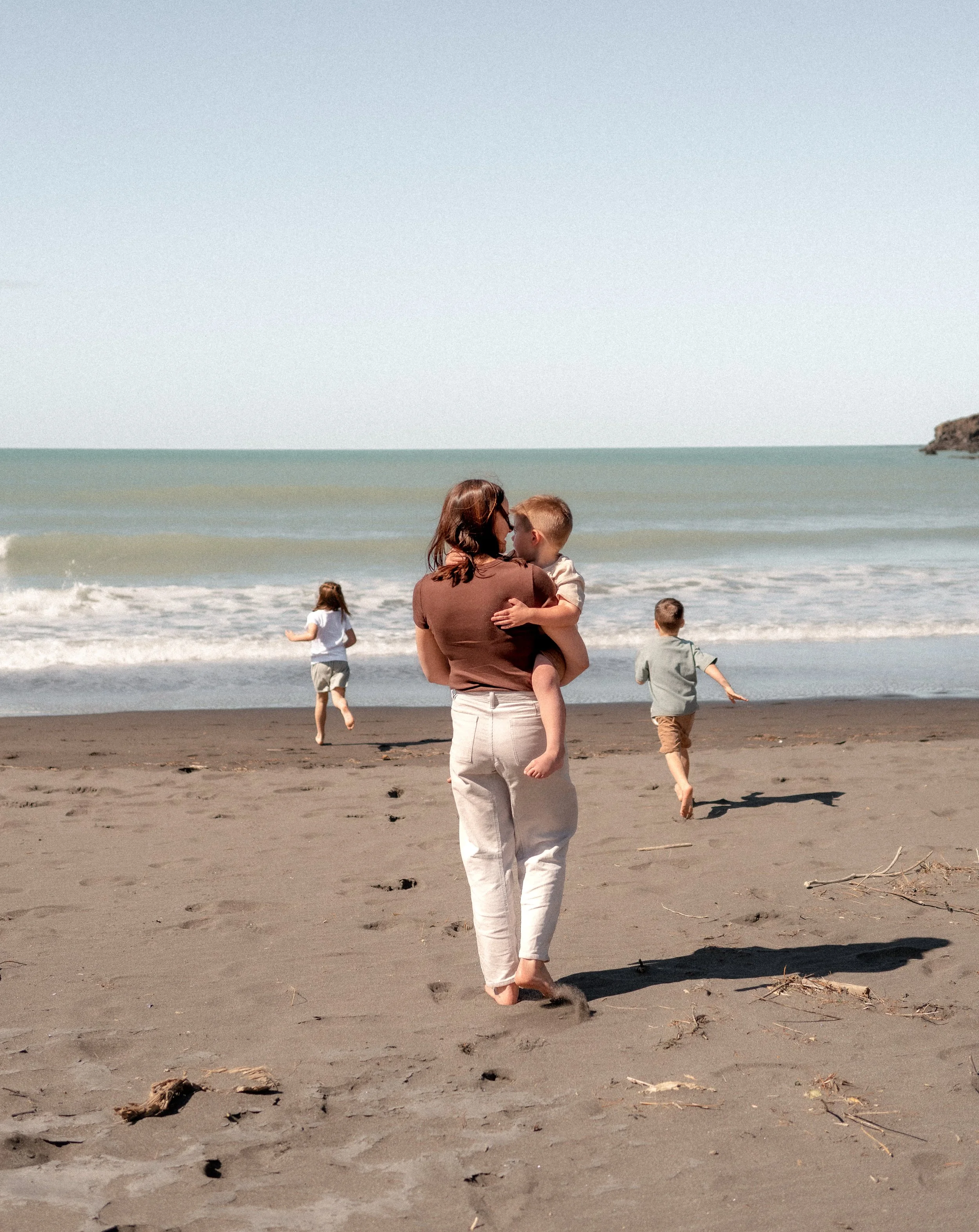 Holistic wellness coach - Alex Ballagh - is walking on a Christchurch beach holding her son and her other 2 children are running towards the ocean.