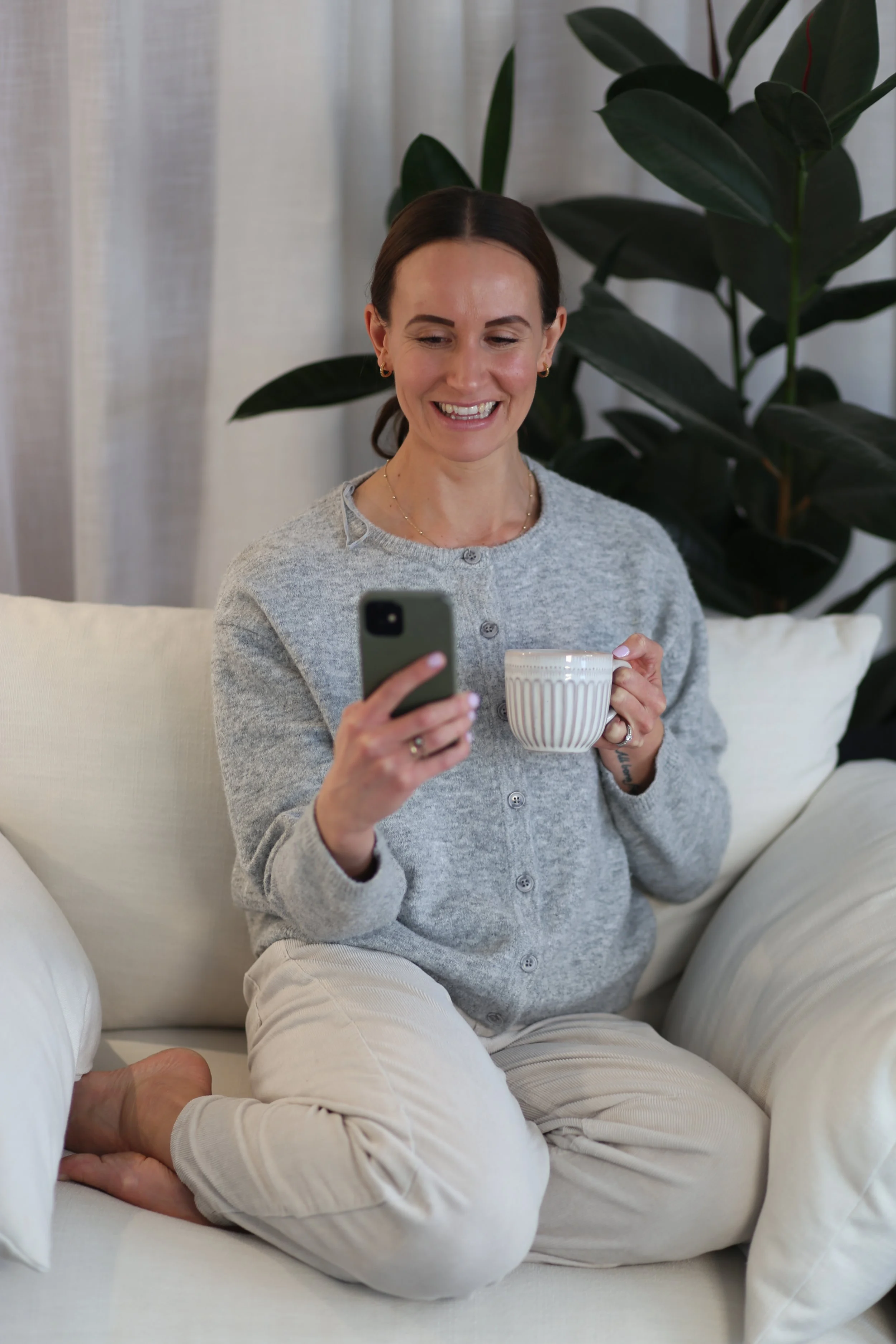 Holistic Wellness Coach - Alex Ballagh - is sitting on a white couch with a cup of tea and looking at her iPhone.