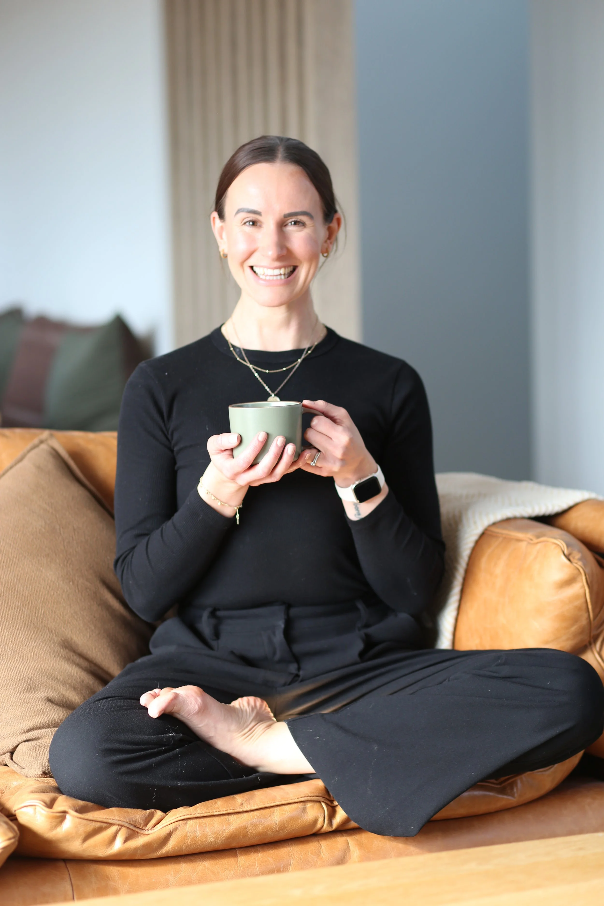 Holistic wellness coach - Alex Ballagh - is sitting cross-legged on a brown leather coach, wearing all black, and smiling at the camera whilst holding a cup of tea in both hands.
