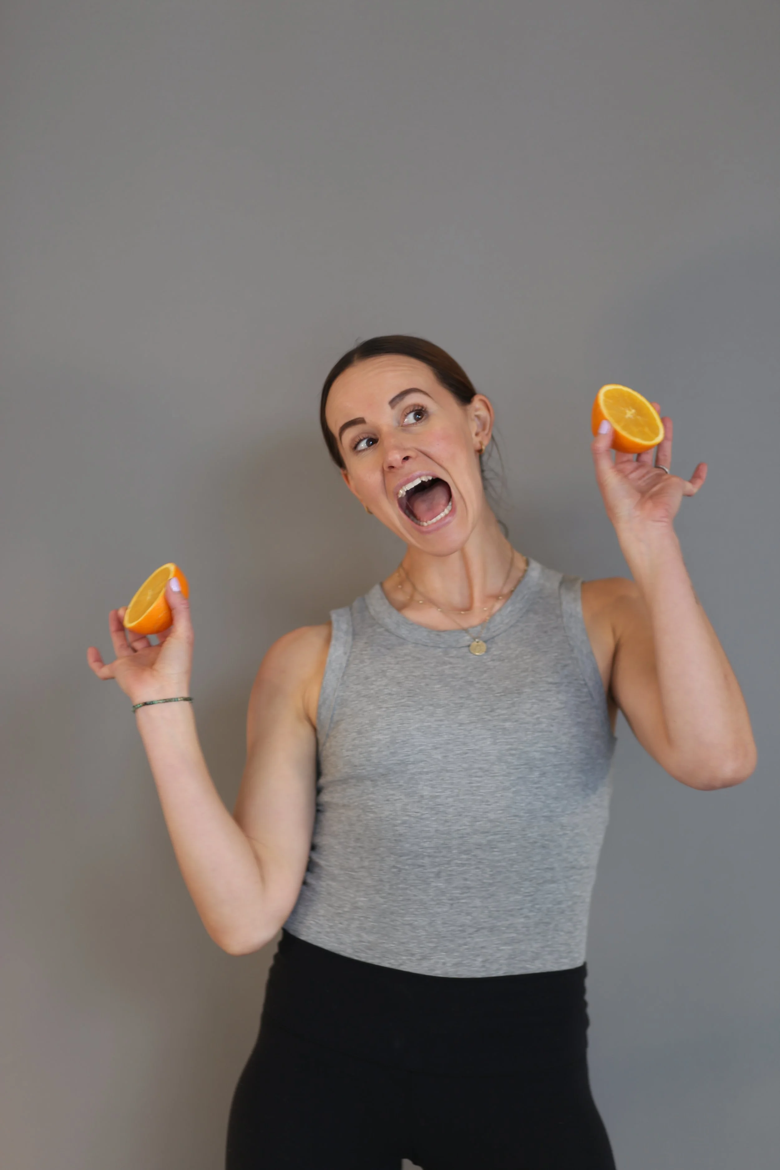 Holistic wellness coach - Alex Ballagh - is smiling cheekily at the camera holding up two halves of an orange in both hands and wearing a grey and black yoga outfit.