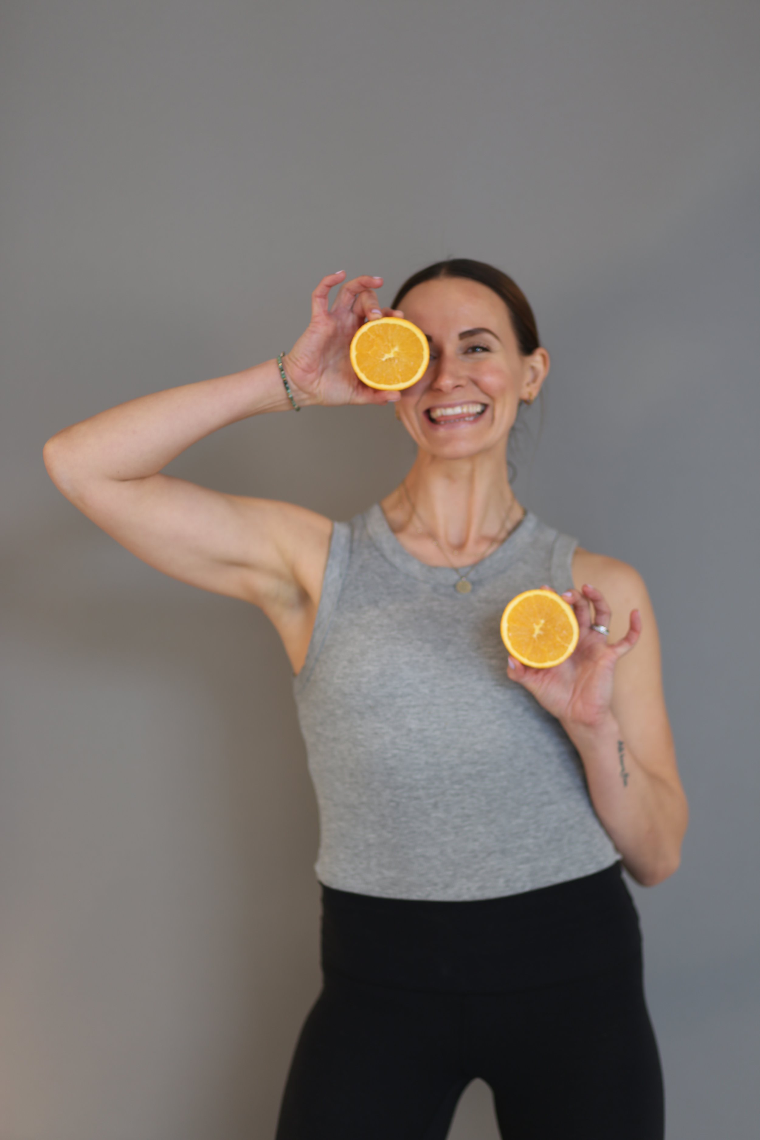 Holistic wellness coach - Alex Ballagh - is smiling and holding 2 halves of an orange, one right in front of her face. She is wearing a grey and black yoga outfit.