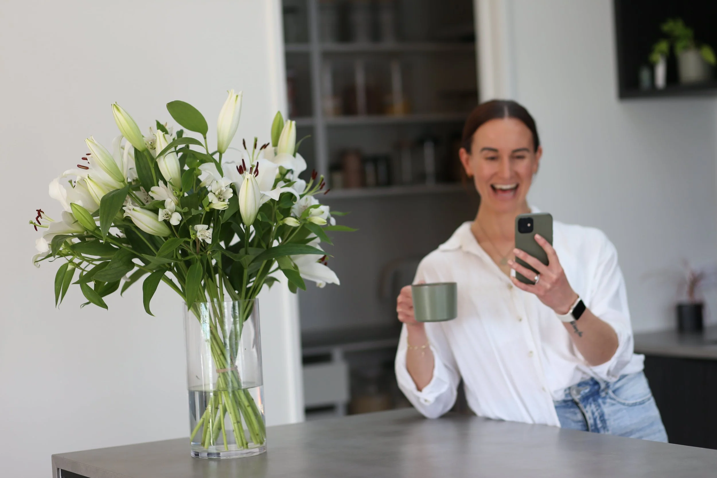 Holistic wellness coach - Alex Ballagh - is sitting at her kitchen bench, holding a green mug of tea and is laughing whilst looking at her phone.