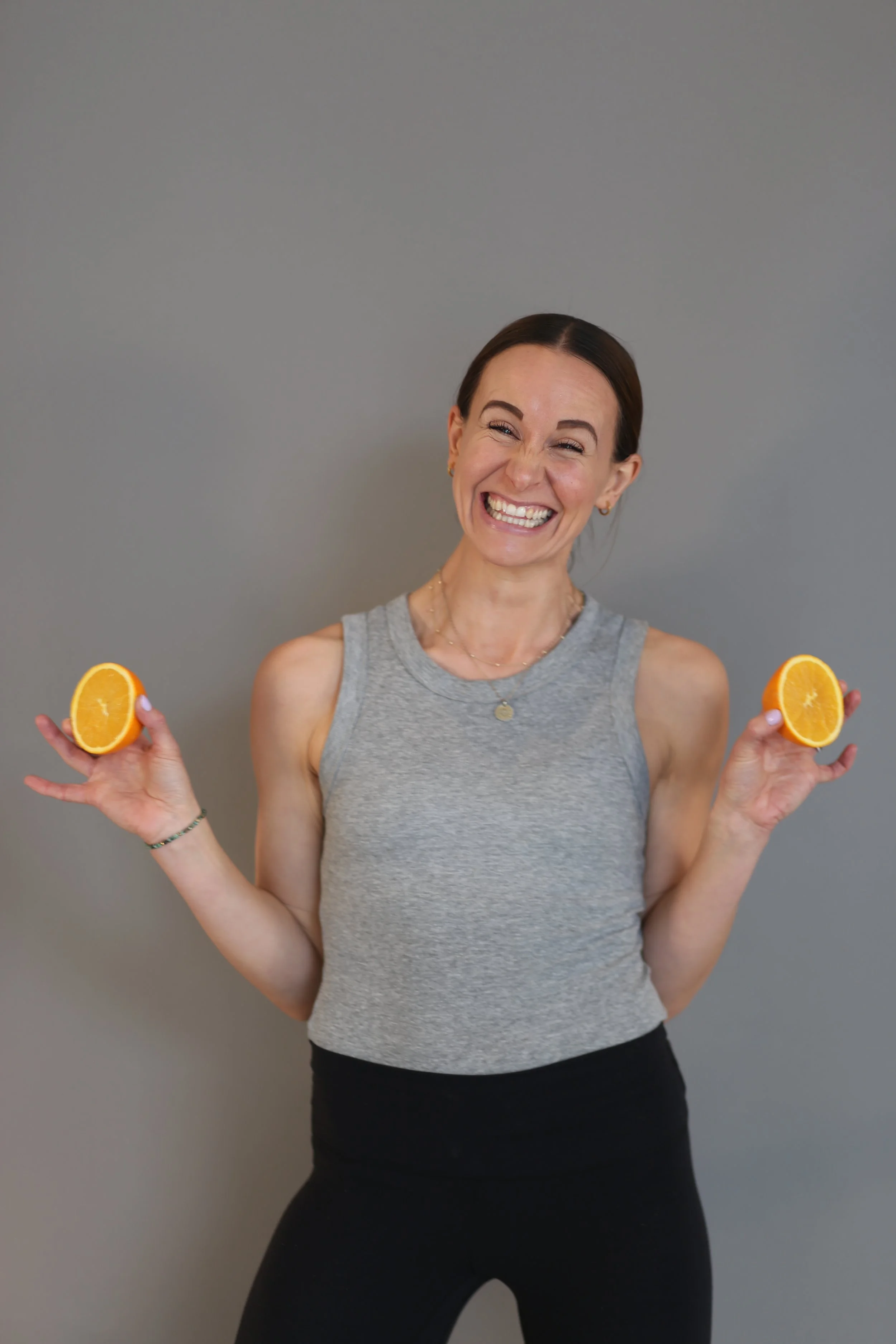 Holistic wellness coach - Alex Ballagh - is smiling cheekily at the camera holding up two halves of an orange in both hands and wearing a grey and black yoga outfit.