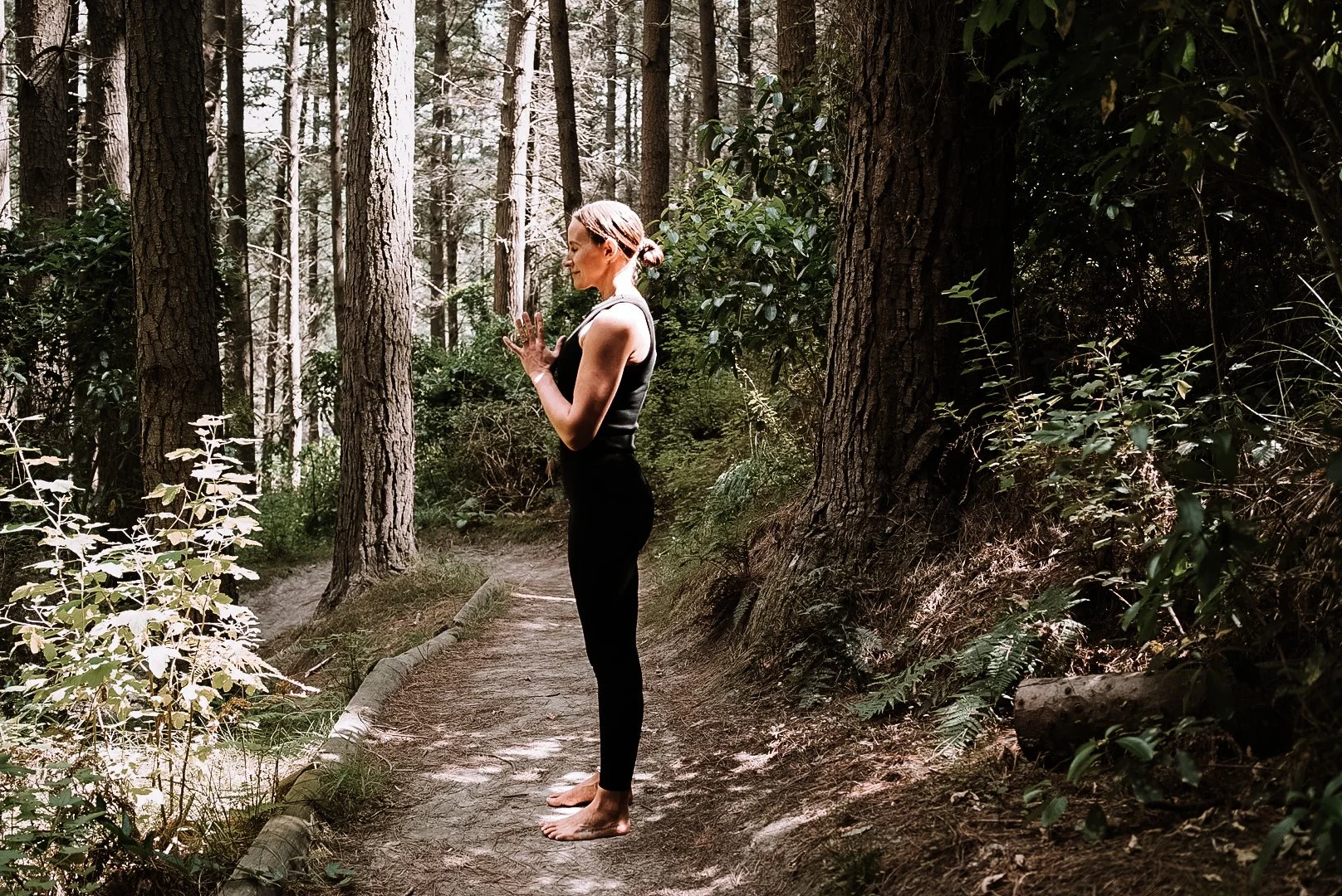 Christchurch holistic wellness coach - Alex Ballagh - is standing in the middle of a pine tree forest in a peaceful yoga pose, holding her hands together.