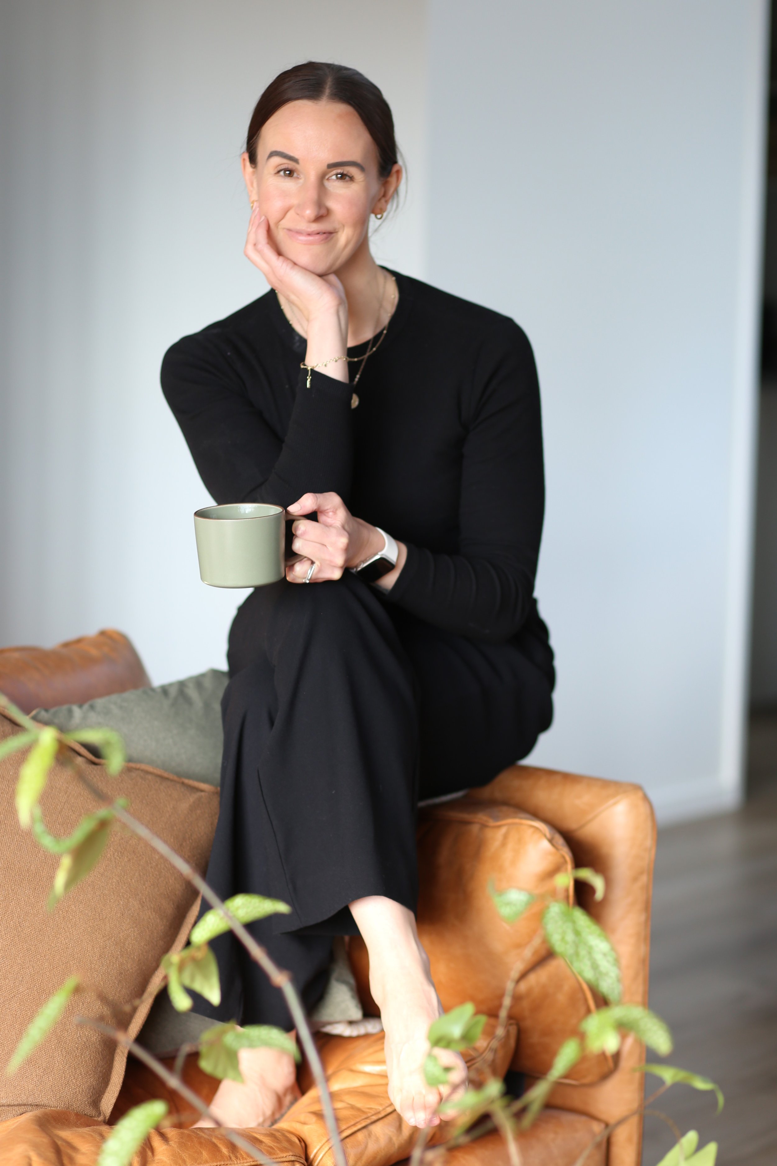 Christchurch holistic wellness coach - Alex Ballagh - is sitting on the arm of a brown leather couch with her leg crossed, and holding a cup of tea.
