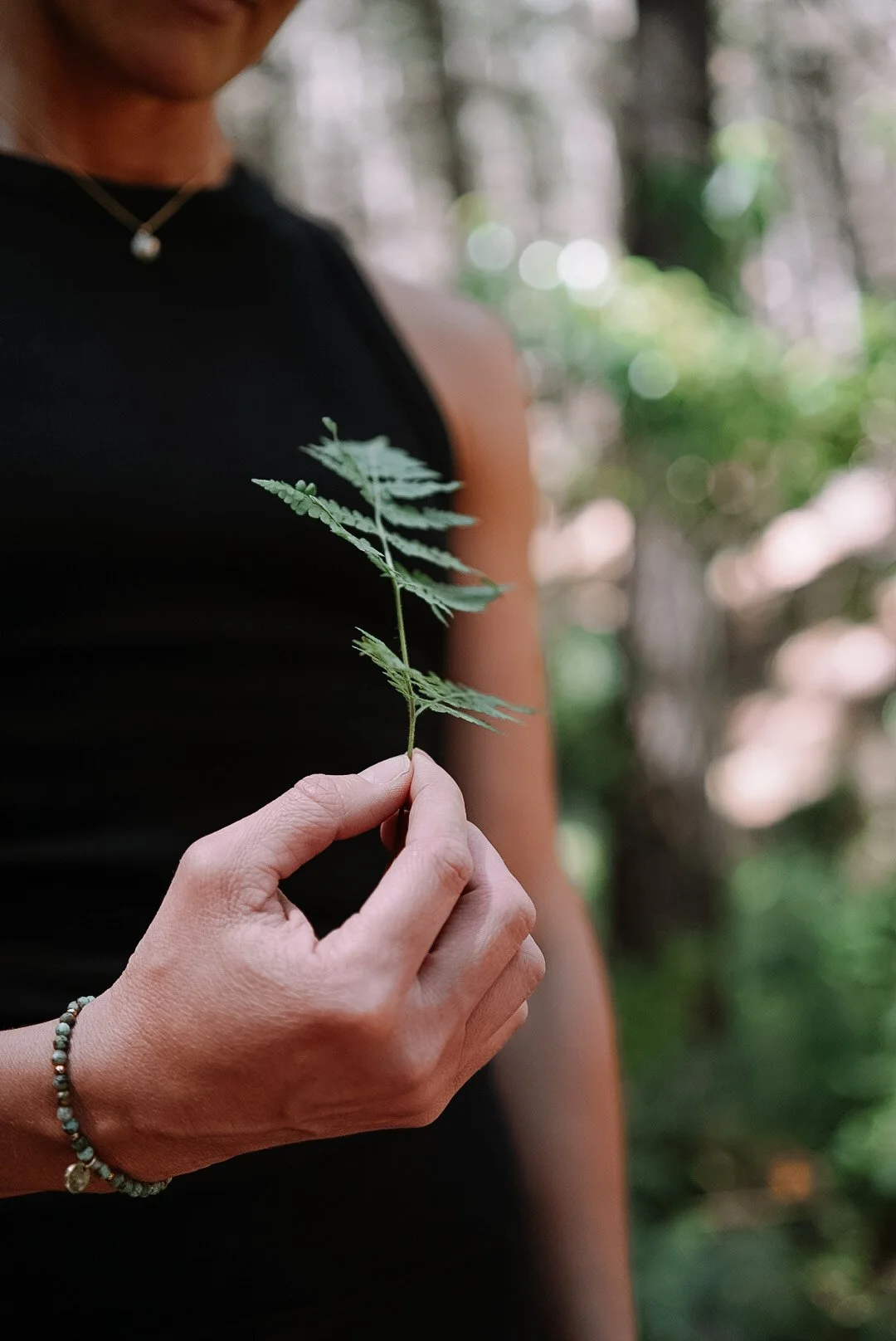 Holistic wellness coach - Alex Ballagh - is standing in the forest and holding up a fern leaf close to the camera.