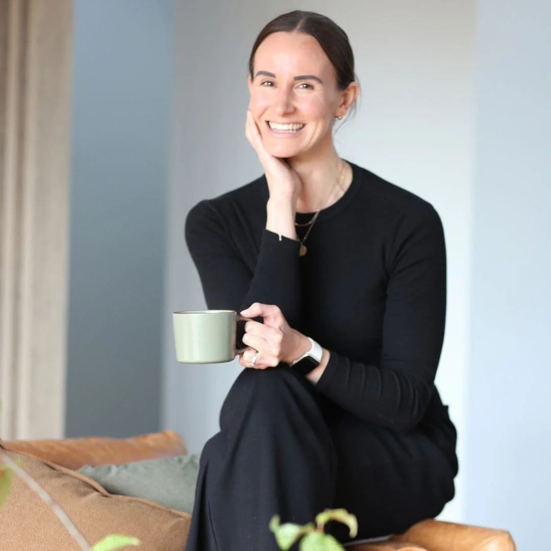 Holistic Wellness Coach, Alex Ballagh, is sitting on the arm of a brown leather couch smiling and holding a cup of tea.