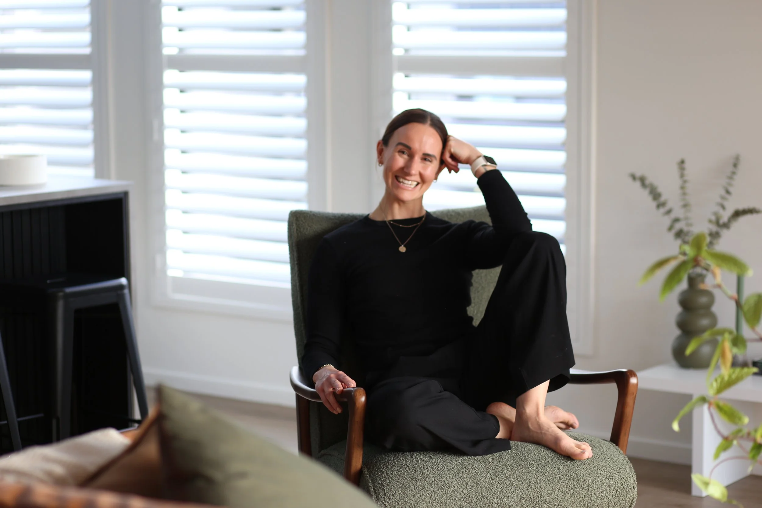 Holistic wellness coach - Alex Ballagh - is sitting on a green chair smiling at the camera, holding her head against her hand.