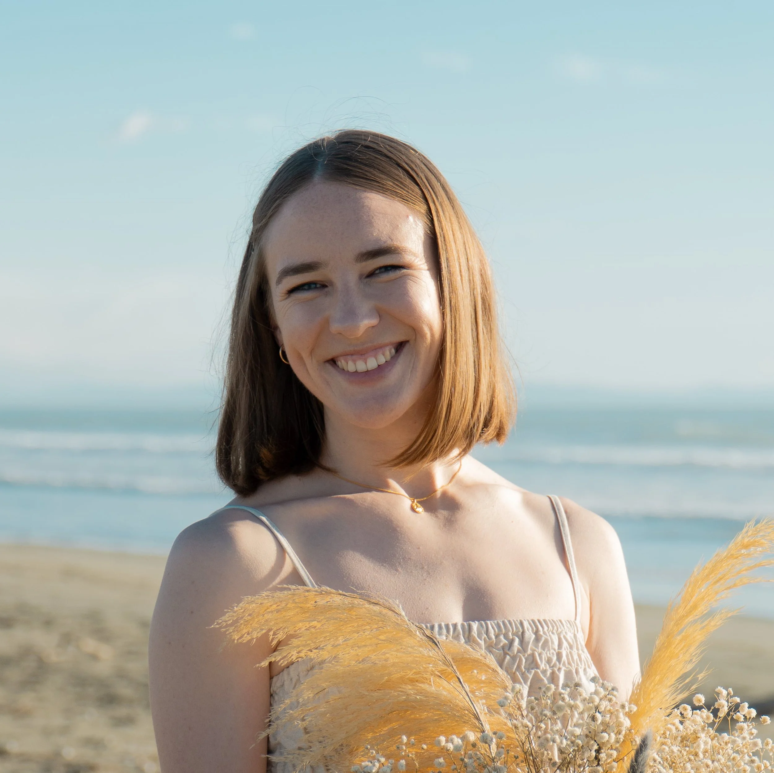 Headshot of Alex's previous coaching client who is smiling at the camera, standing on the beach and wearing white dress and holding flowers.