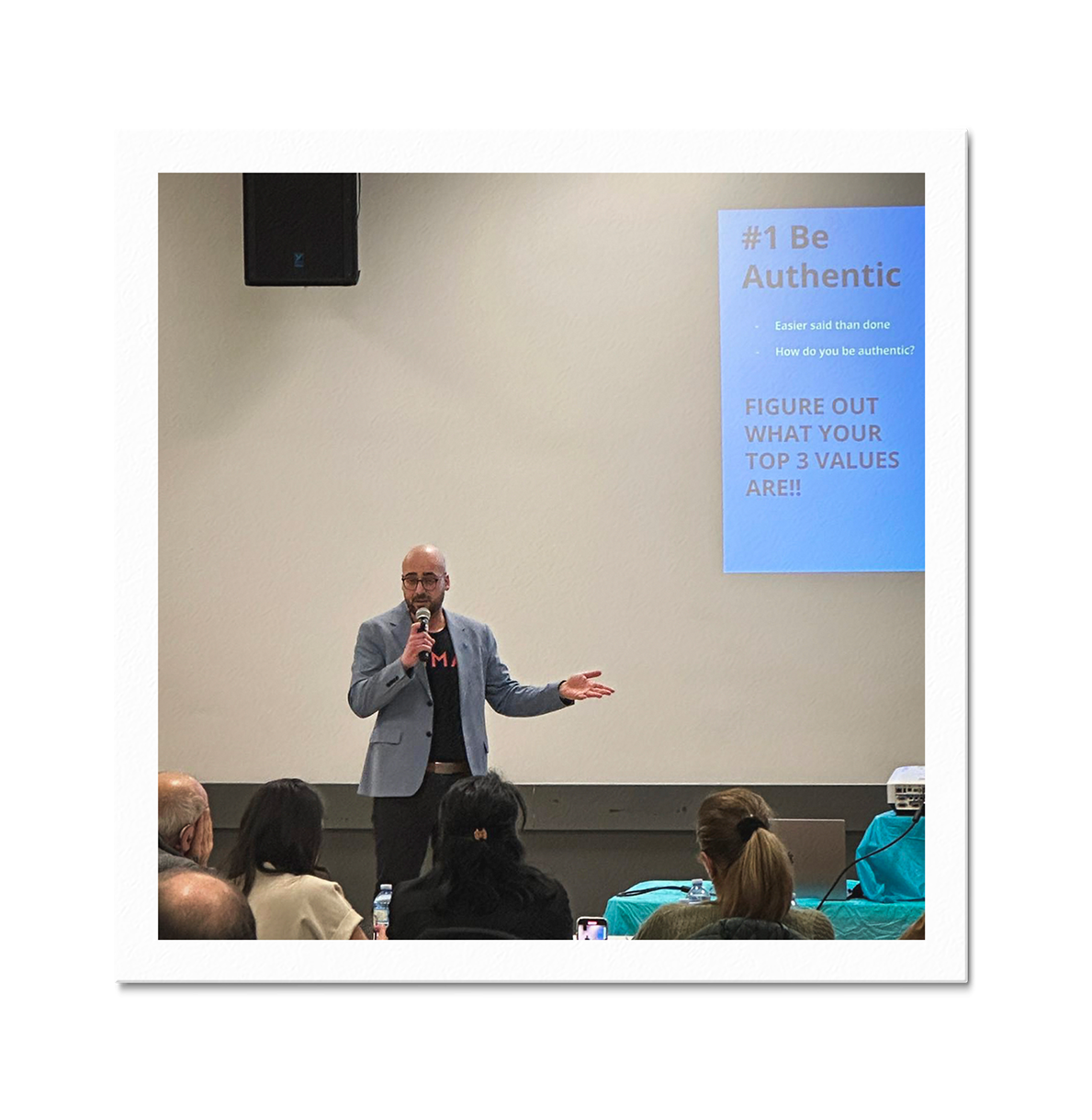 A man in a gray blazer giving a presentation to an audience, with a blue slide on the wall that reads '#1 Be Authentic' and 'Figure out what your top 3 values are!!'