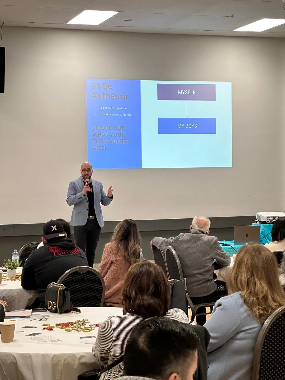 A man in a light gray blazer giving a presentation at a conference or workshop, with a slide projected behind him titled '#1 Be Authentic' that discusses values and being true to oneself and one's children.