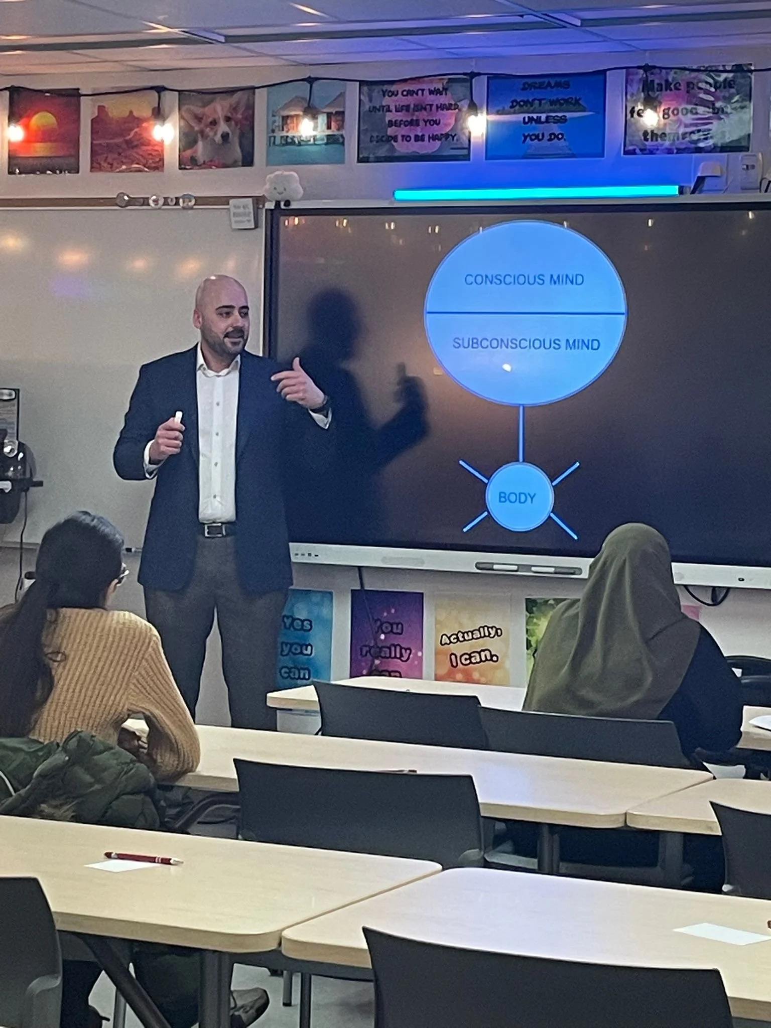 A man giving a presentation on the conscious and subconscious mind in a classroom. There are students seated at desks, and a large screen displaying a diagram of the mind's structure.