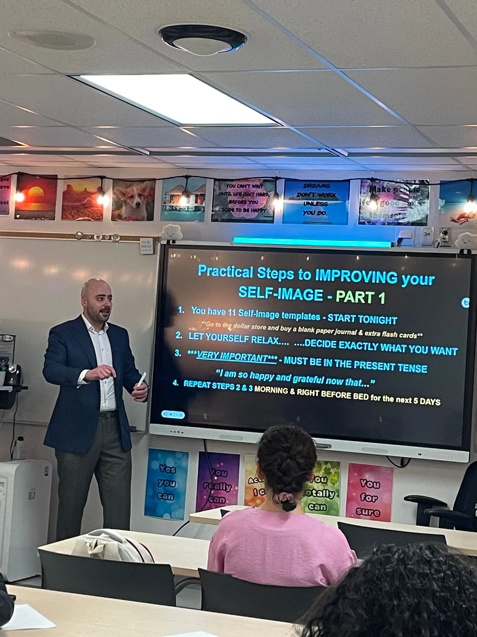 A man giving a presentation in front of a large screen with text about improving self-image, in a classroom or meeting room setting. There are colorful posters and flags on the wall behind him.