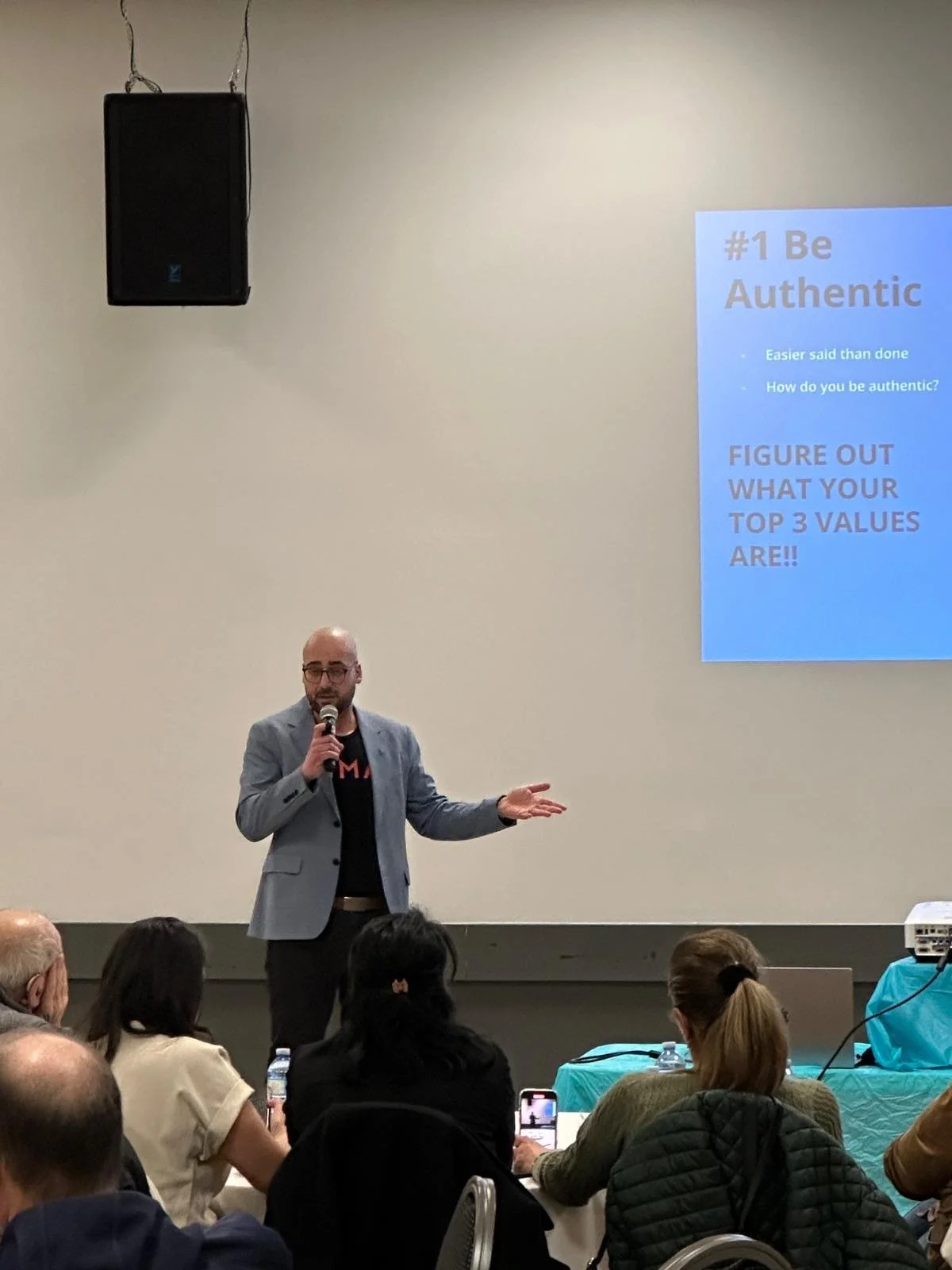 A man with glasses and a gray blazer is speaking into a microphone while gesturing with his left hand in front of a seated audience. A large screen to his right displays a presentation slide titled '#1 Be Authentic' with additional text.