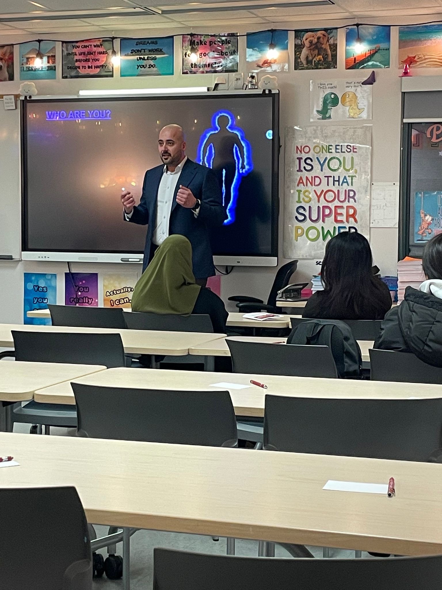 A man giving a presentation in a classroom with students seated at desks. The presentation slide shows a silhouette of a person with the text 'WHO ARE YOU?'. Classroom decorations include motivational posters and colorful artwork on the walls.