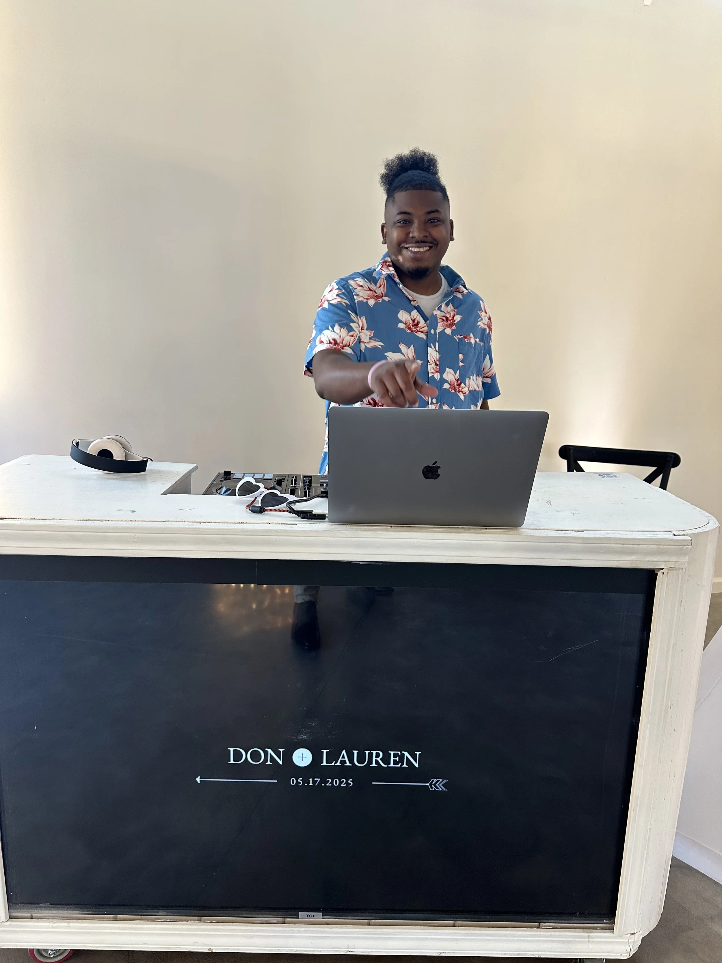 A young man with dark curly hair tied up in a puff, wearing a blue floral shirt, stands behind a white wooden table with a laptop, DJ equipment, and headphones on it. The table has a black screen display with the names 'Don + Lauren' and the date 05.17.2025, with decorative arrows.