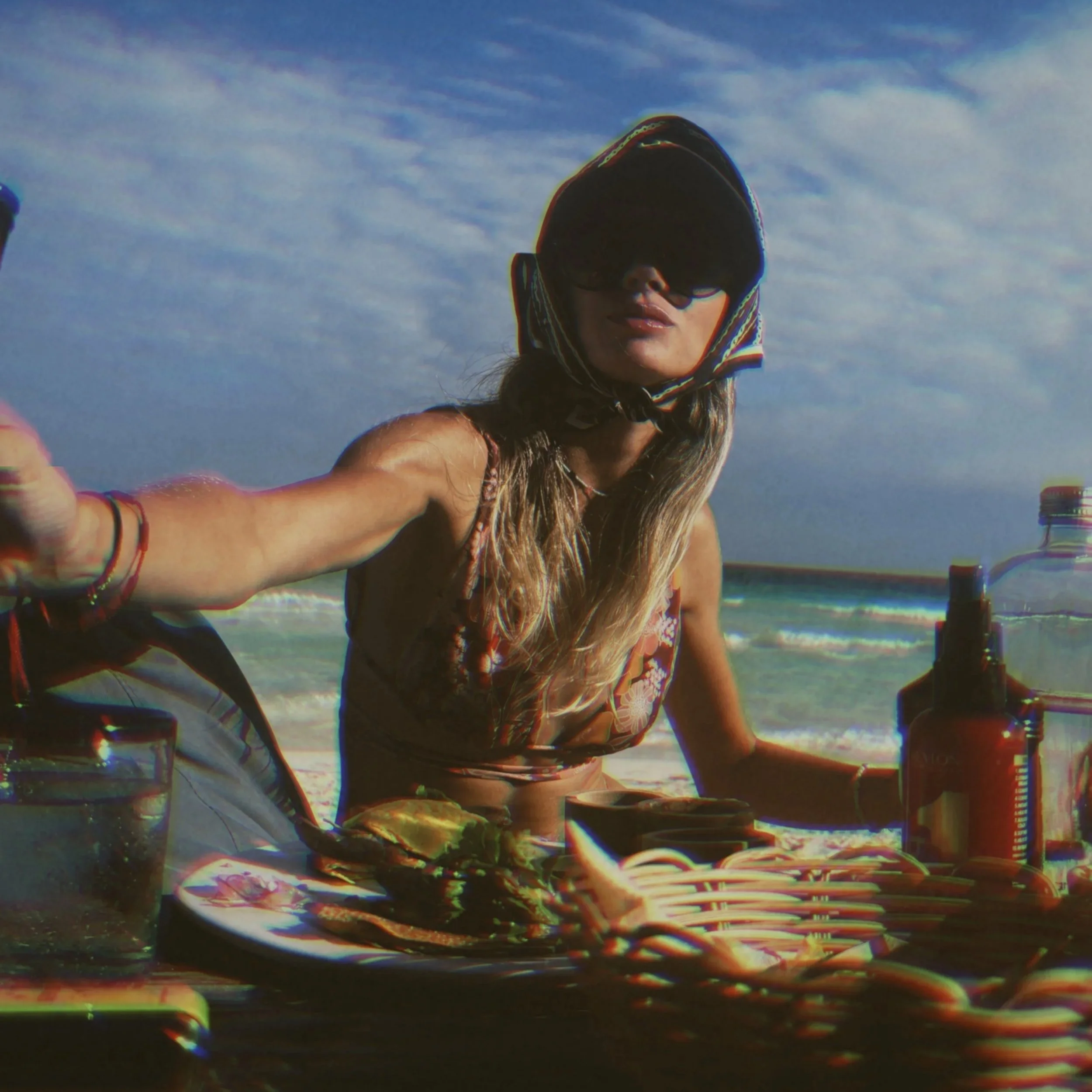 Woman sitting at a table on the beach, wearing sunglasses and a hood, with ocean and sky in the background.