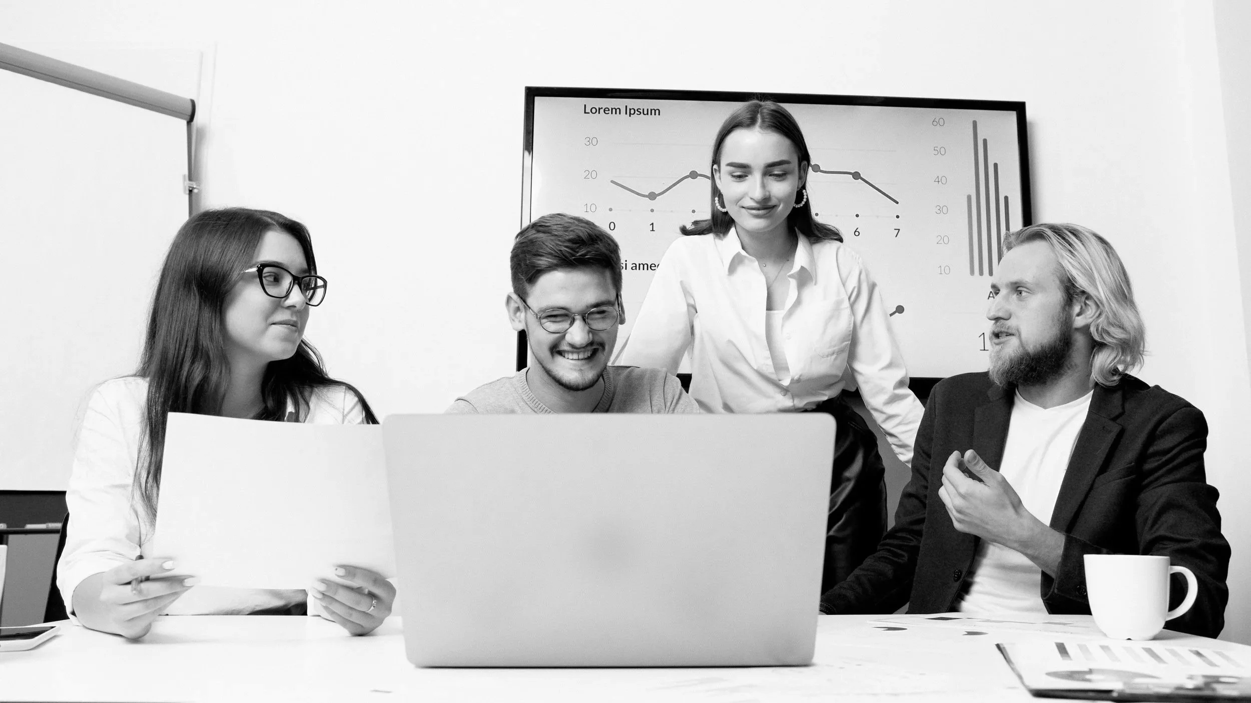 Four people in a business meeting looking at a laptop in a conference room with charts on a screen in the background.