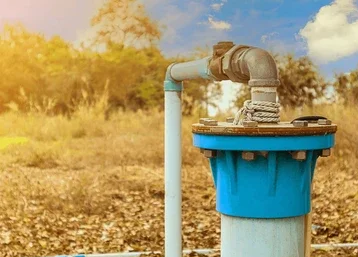 A blue water meter with a pipe connected to it outdoors in a natural setting.
