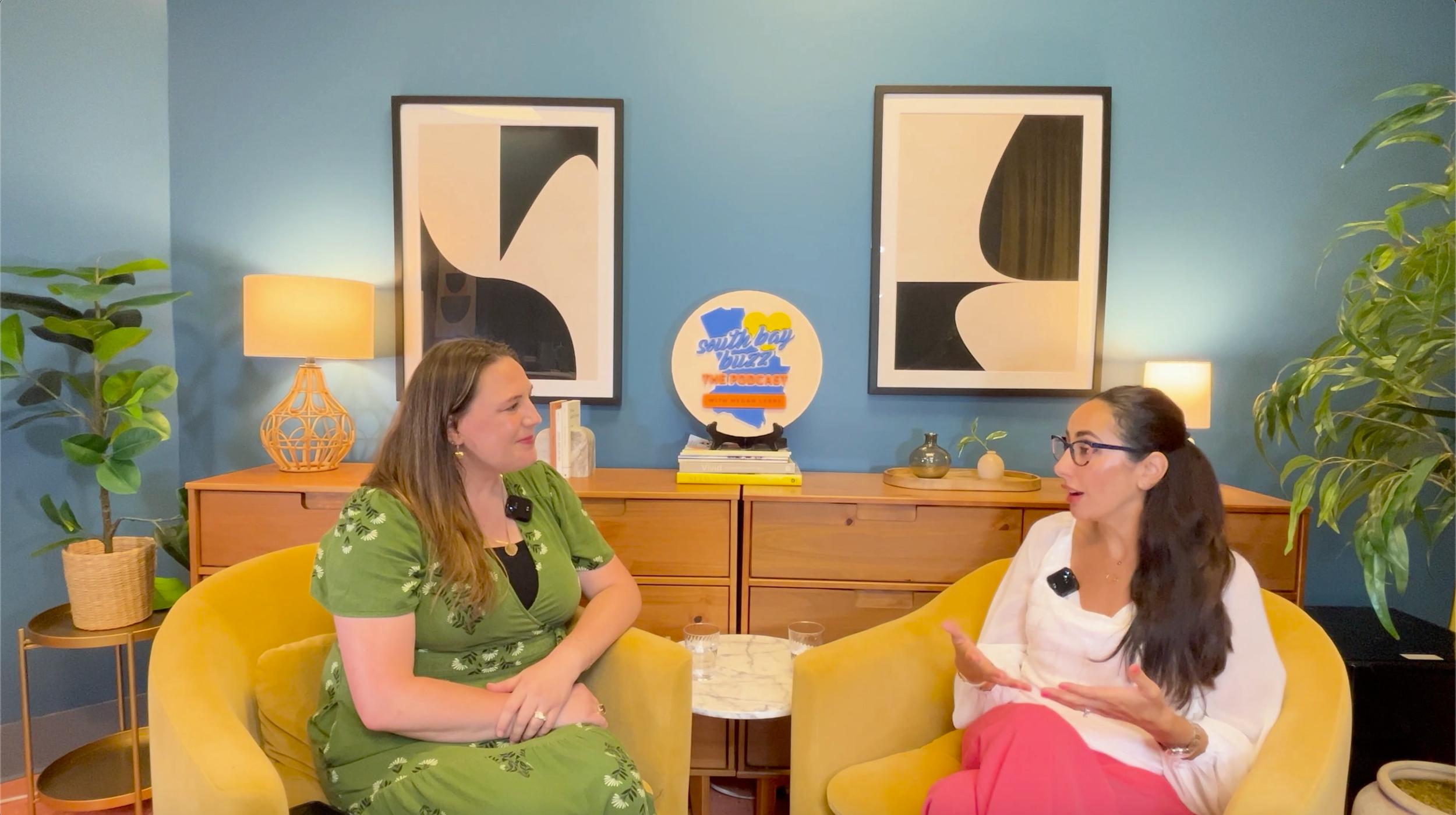 two women talking in yellow chairs in a blue room with two green ferns in corner.