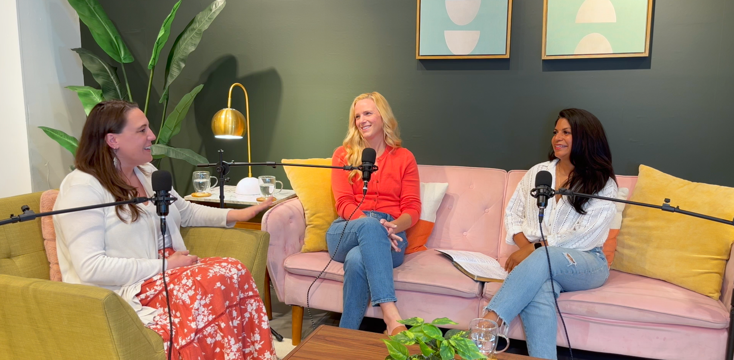1 women in a yellow chair and 2 women on a pink couch talking into microphones on a podcast smiling at each other