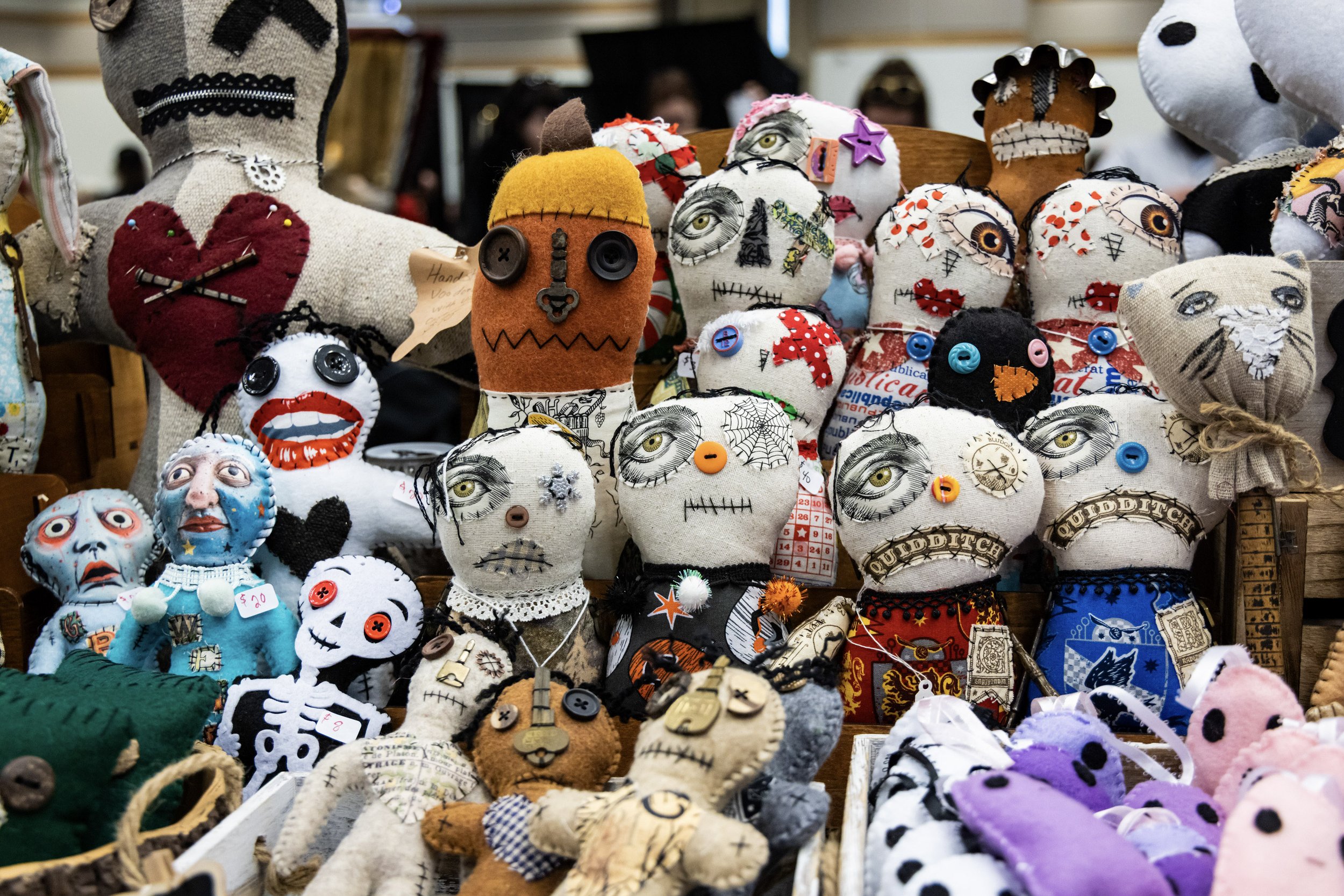 A variety of handmade horror-themed cloth and felt dolls with sewn features and button eyes, including skull faces, monsters, and skeletons, displayed on a table at a craft market.