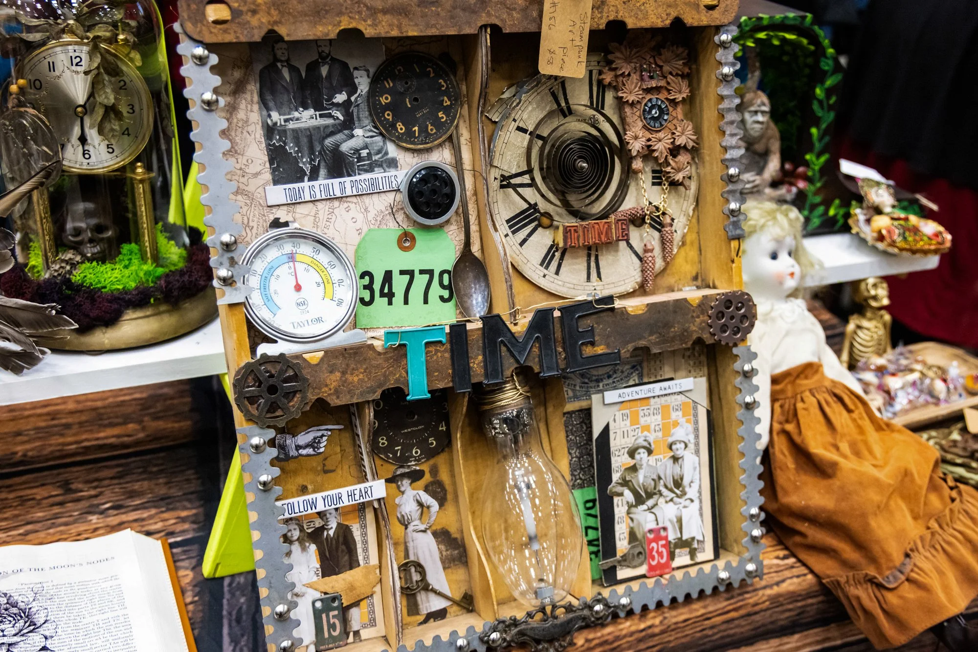 A decorative collage of vintage clocks, black and white photos, and assorted trinkets on a wooden board. Includes clocks showing different times, a black and white photo of people in vintage clothing, and various small objects like a spoon, gears, and a green tag with the number 34779.