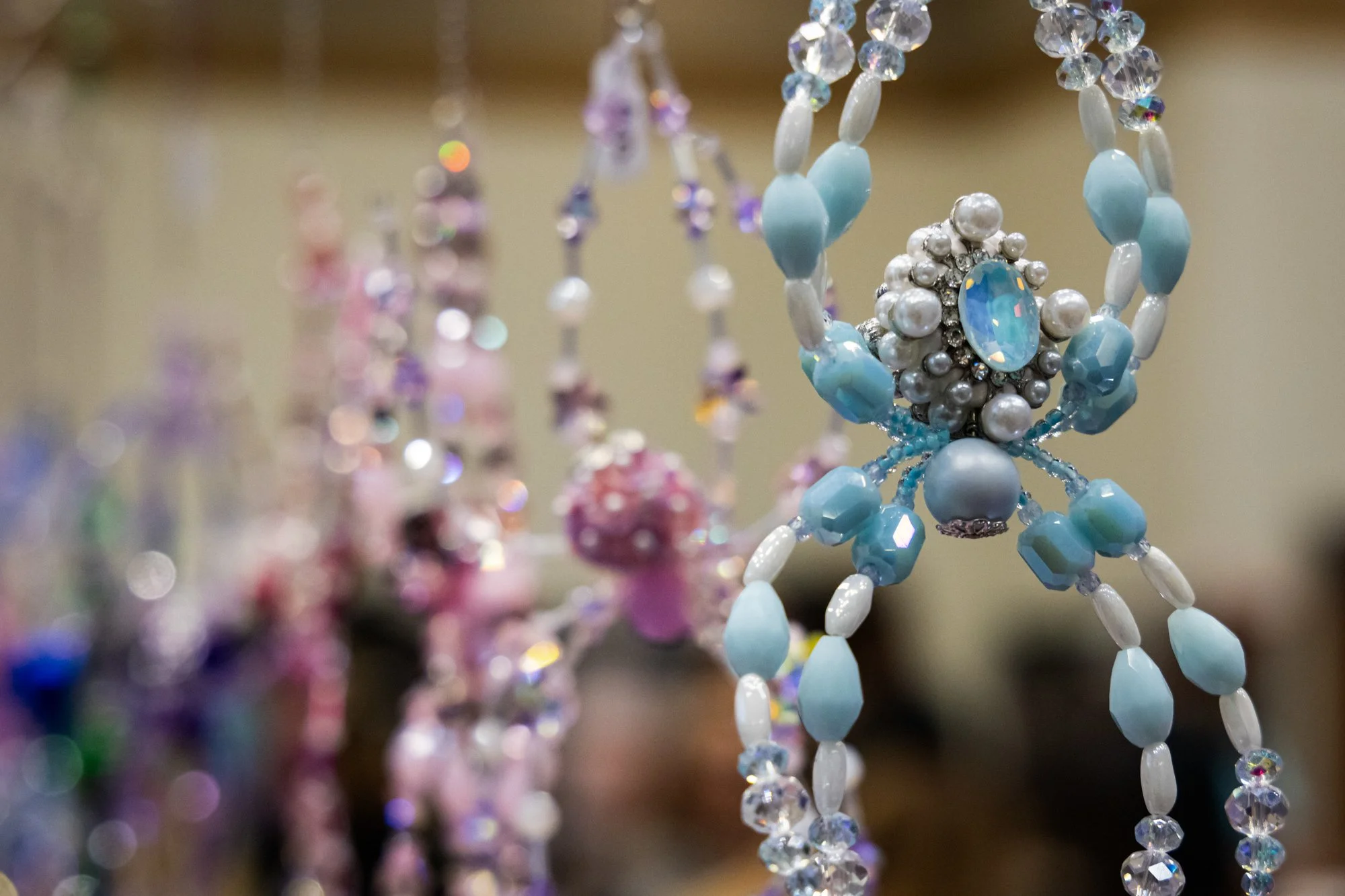 Close-up of beaded jewelry, including a necklace with a large central gem surrounded by pearls and blue beads, amidst other colorful beaded accessories in the background.