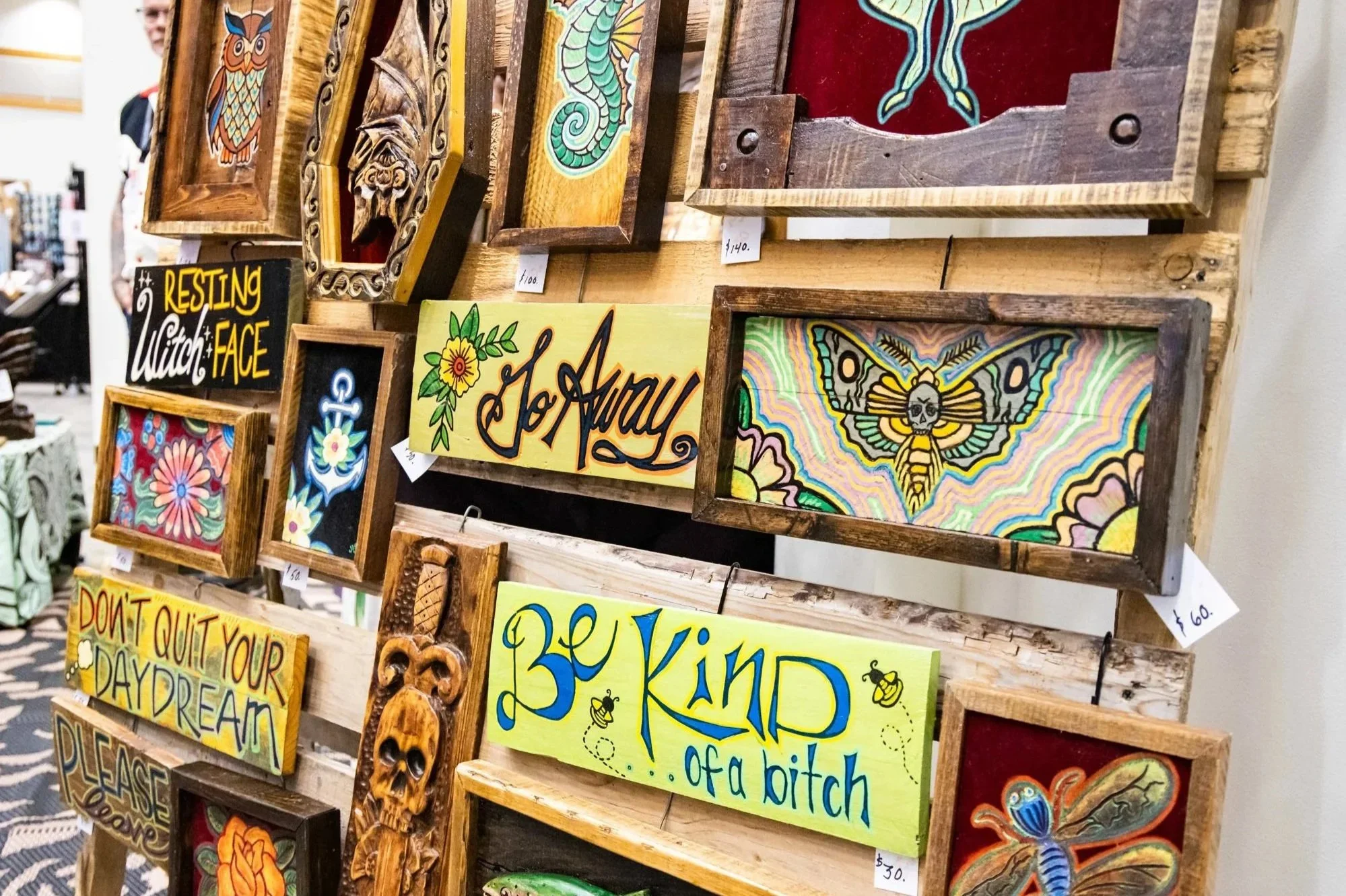 Display of colorful wooden wall art with various designs including an owl, a seahorse, a butterfly, and floral patterns, along with hand-painted signs with inspirational messages, at an indoor market or craft fair.