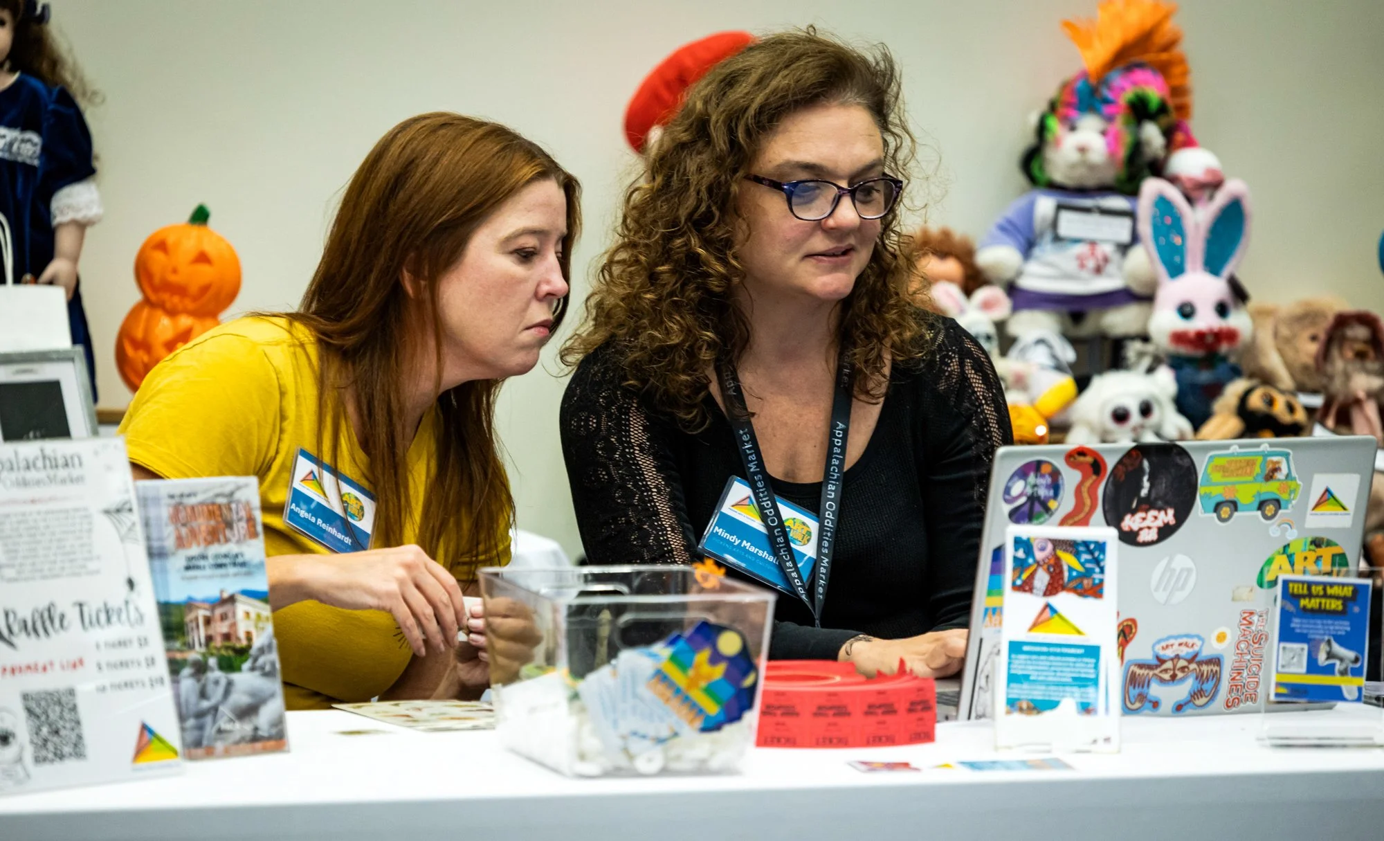 Two women sitting at a table, working on a laptop, surrounded by colorful toys and game tickets, with plush toys and decorations in the background.