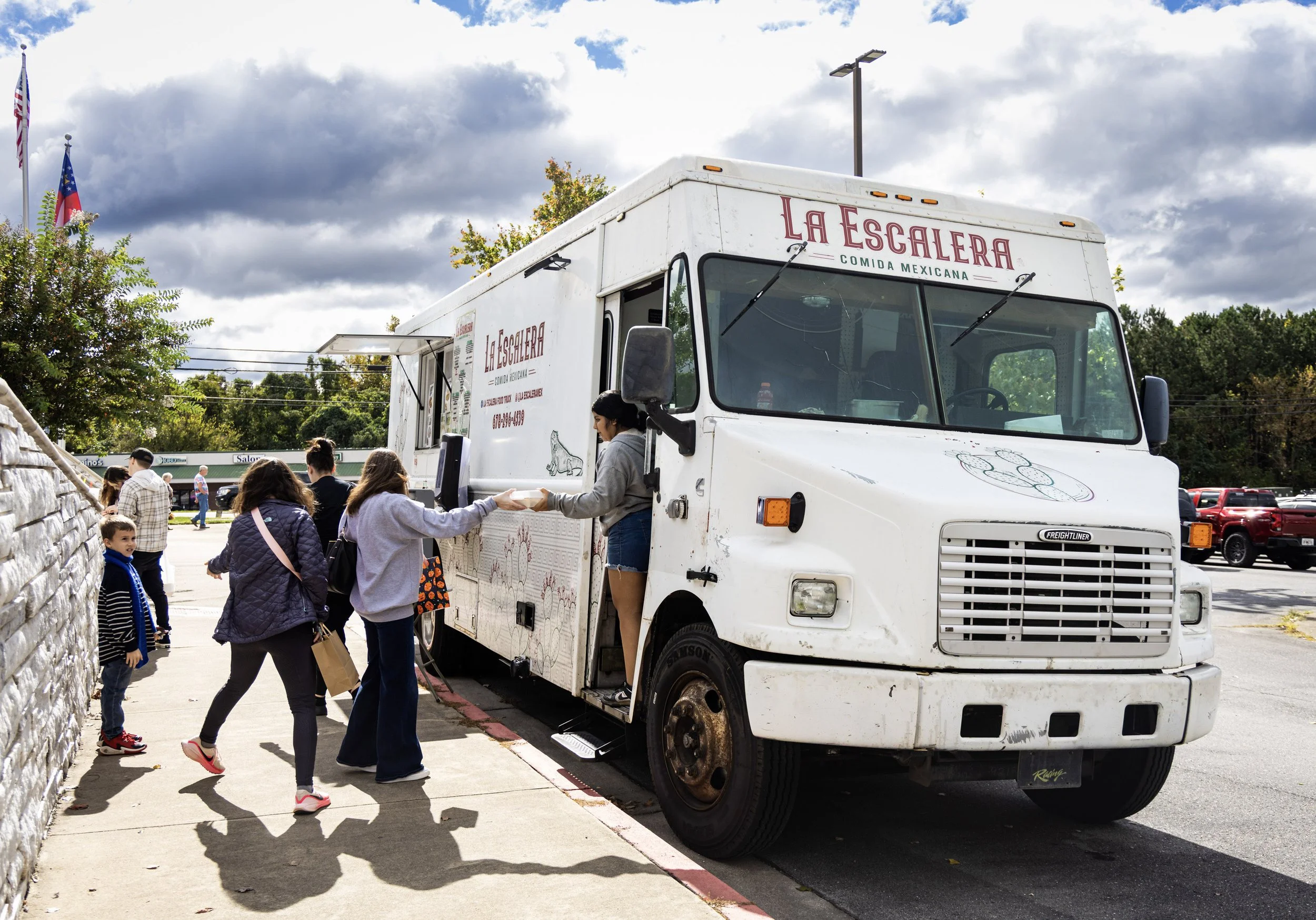 People in line at a food truck named La Escalera serving Mexican food, with a cloudy sky above and flags in the background.
