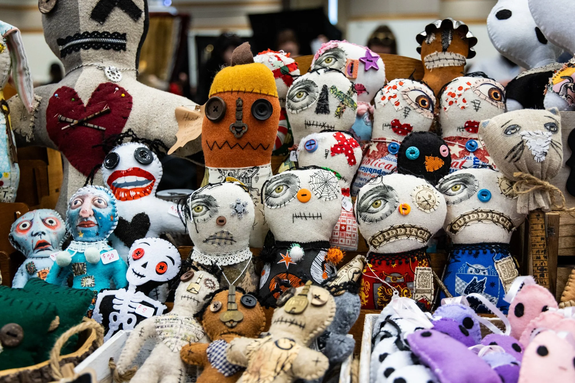 A variety of handmade fabric dolls resembling skeletons, monsters, and skulls are displayed on a table at a craft market. The dolls are decorated with buttons, embroidery, and painted details, some with price tags.