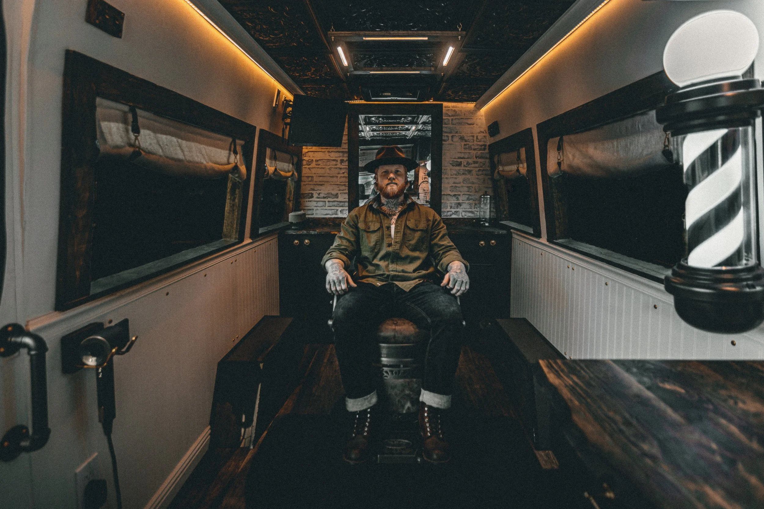 Nomad Mobile Barbershop owner seated in the barber chair inside the van facing the camera with barber pole and tin ceiling, commercial portrait photography by James Brasier